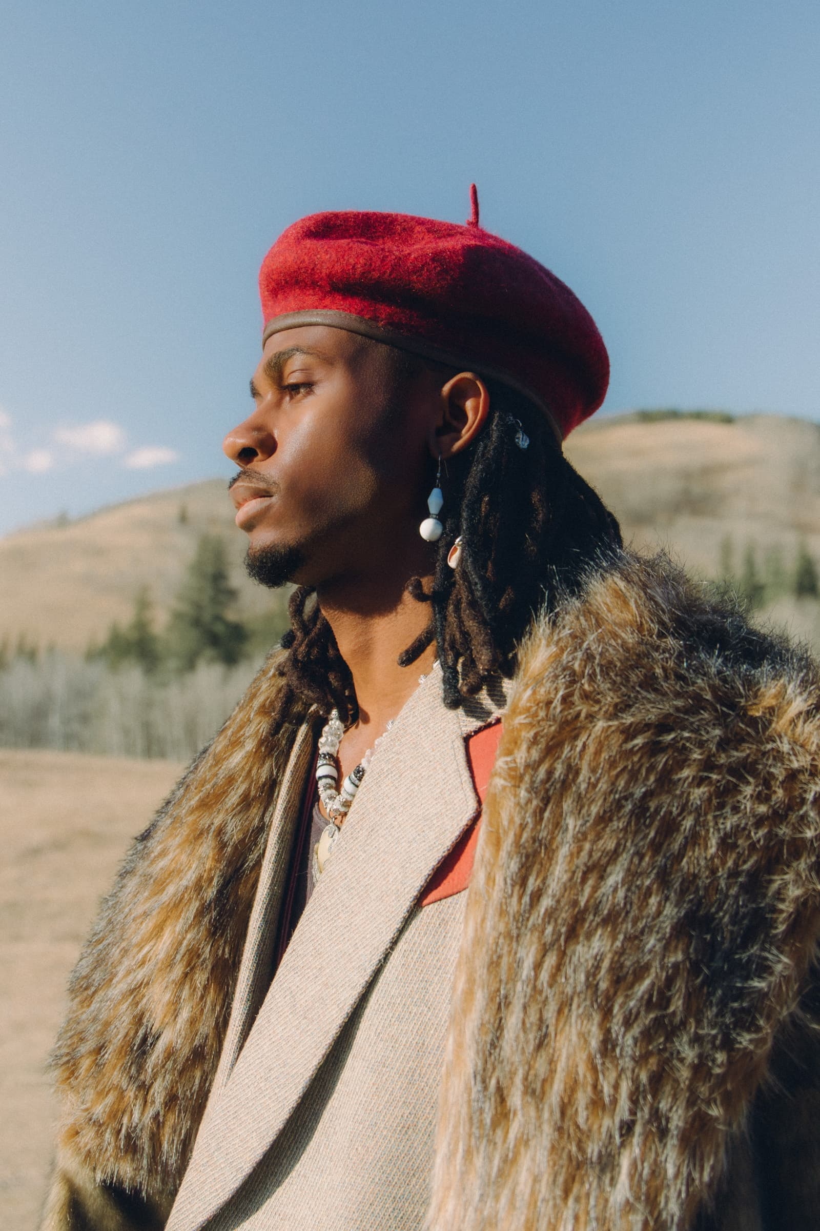 Stylish man in beret, fur coat, and jewelry in scenic outdoor setting.