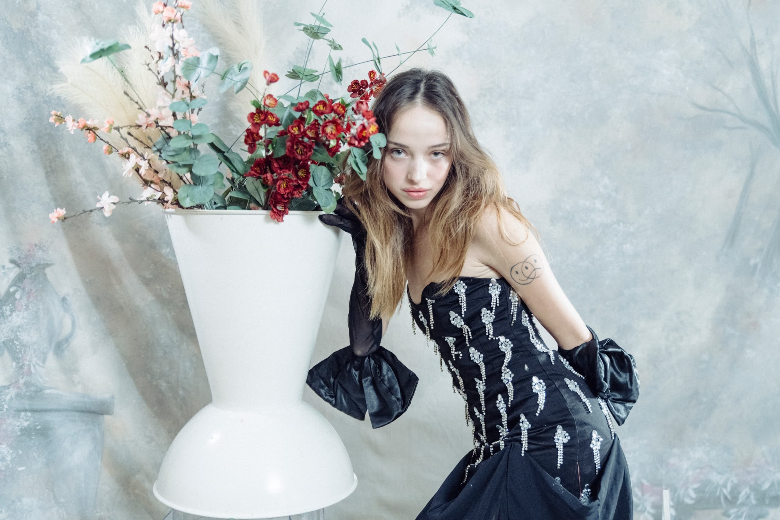 Woman in elegant gown posing by floral vase in artistic setting.