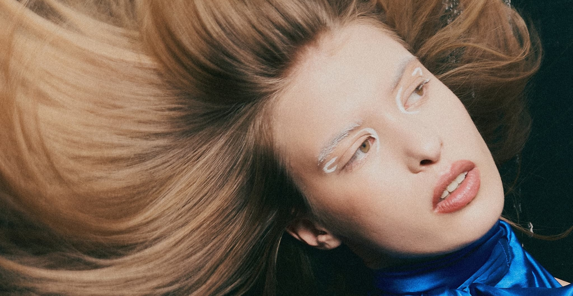 Model with flowing hair and artistic makeup wears a blue blouse, gazing to the side.