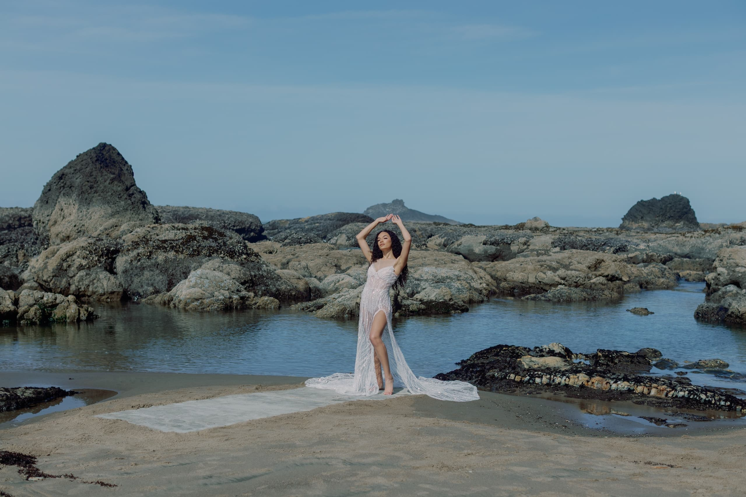 Woman in a flowing dress posing on rocky beach with serene blue skies and water.