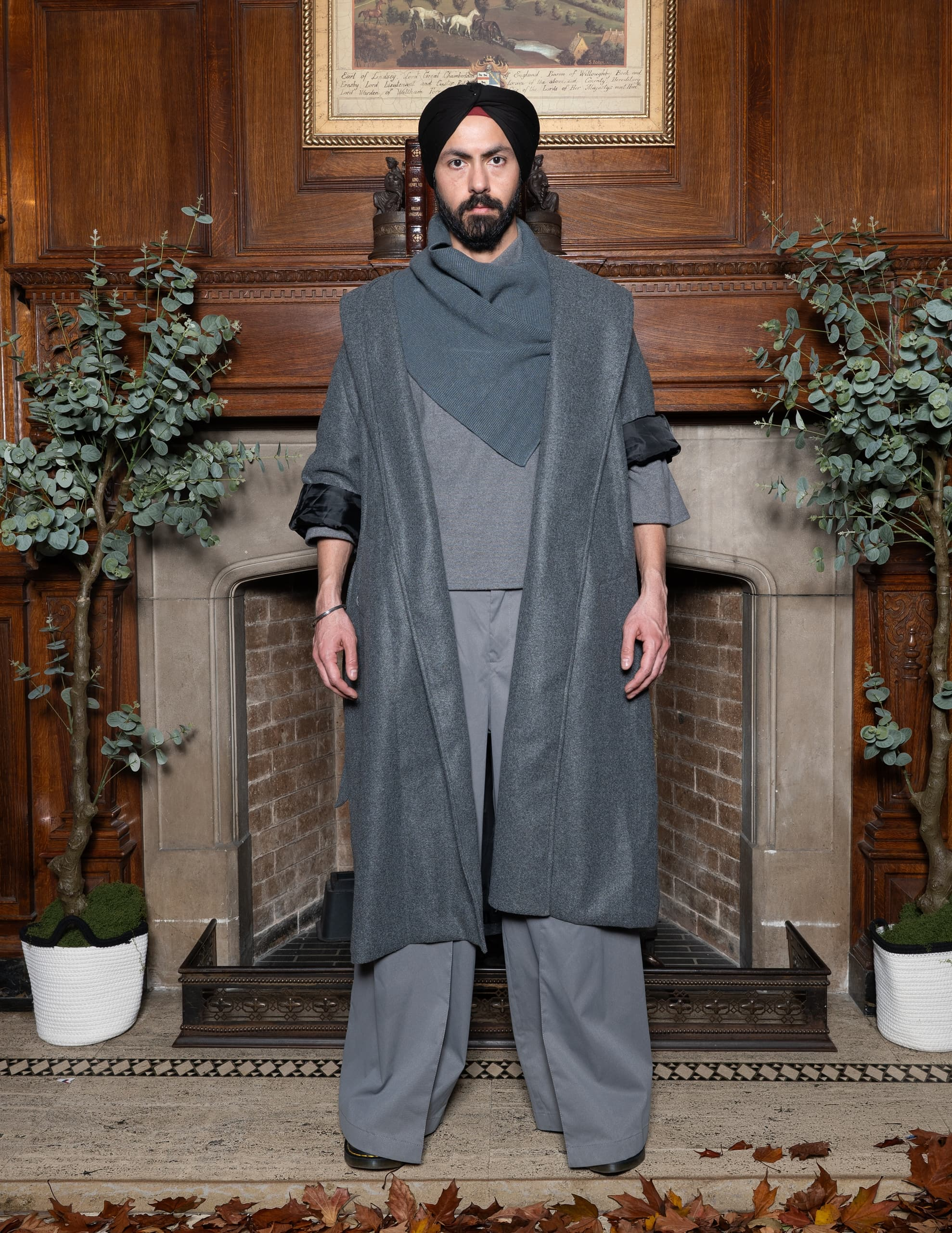 Man in stylish gray outfit standing by a fireplace with a turban and scarf.