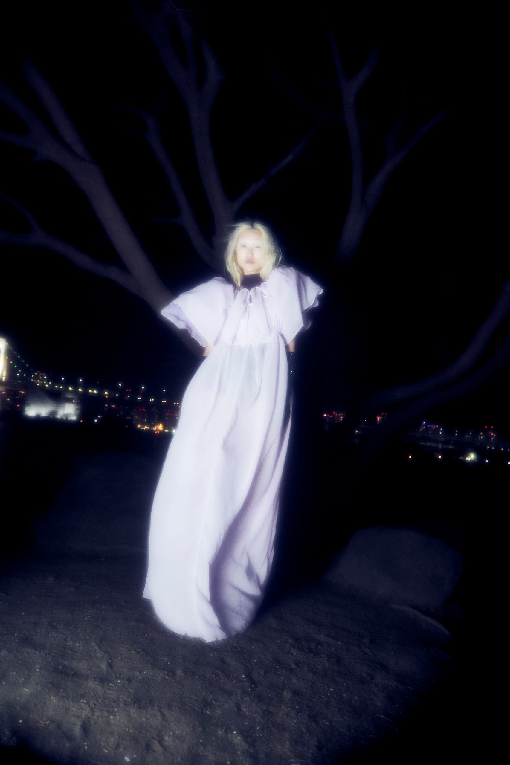 Person in flowing white gown stands at night under a tree with city lights behind.