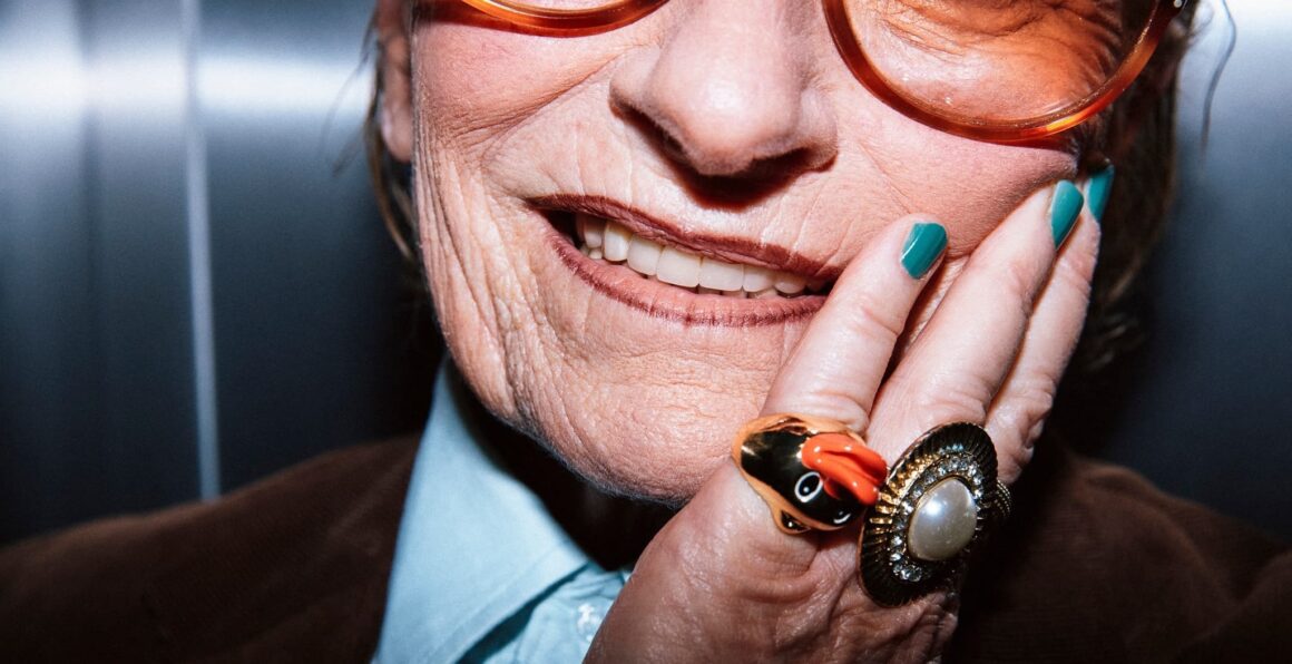 Elderly woman smiling with bold orange glasses and colorful rings.