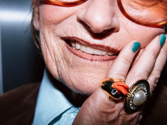 Elderly woman smiling with bold orange glasses and colorful rings.
