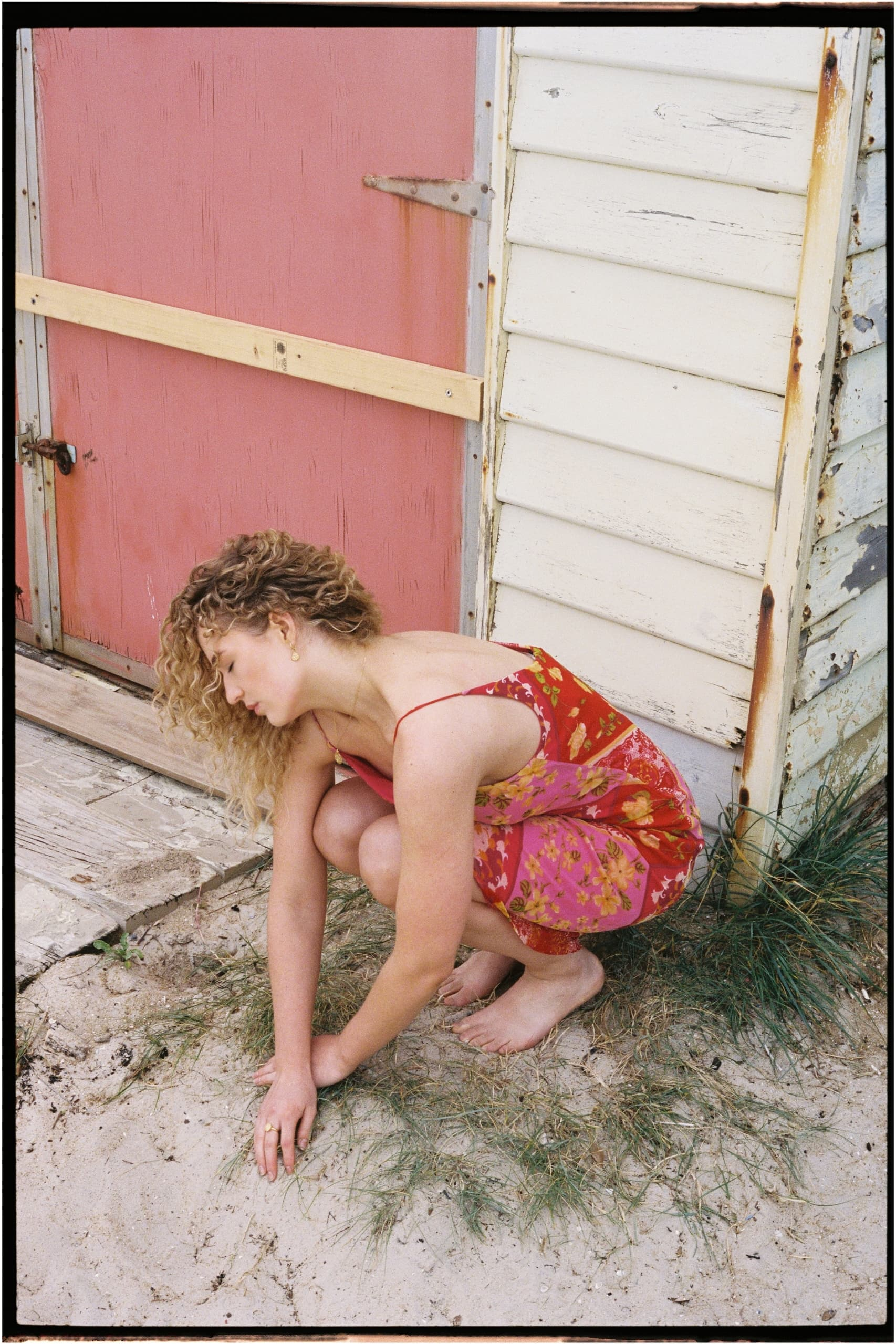 Woman in floral dress crouching on sandy ground by a rustic shed.