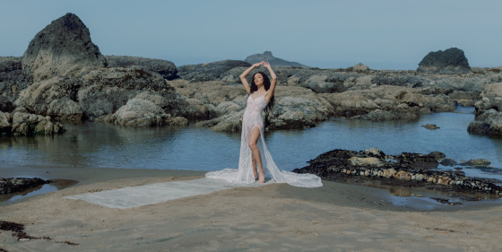 Woman in elegant dress standing on rocky shoreline with arms raised.