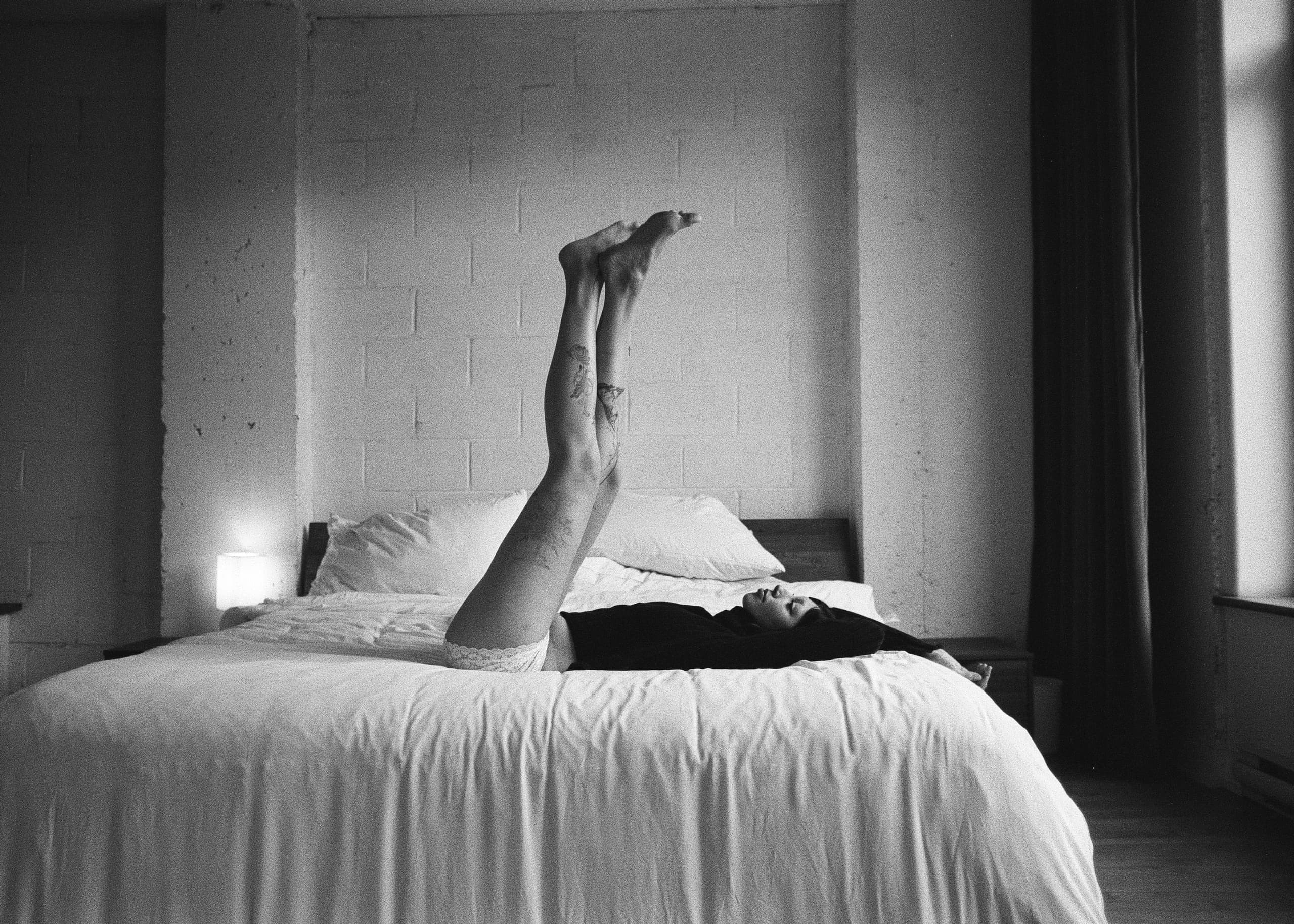 Person relaxing on bed with legs raised, monochrome setting, minimalist bedroom.