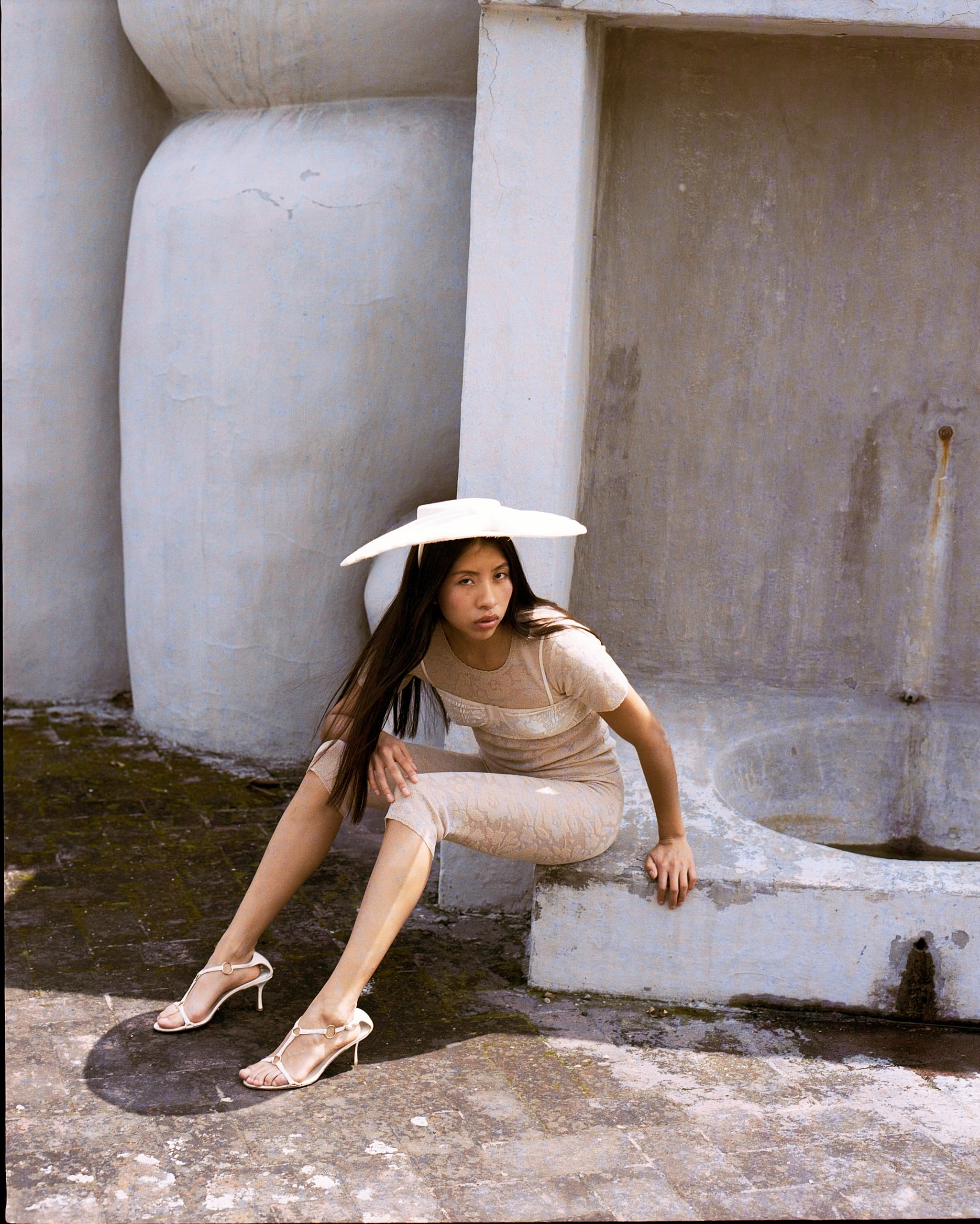 Woman in beige dress and hat sitting by a concrete wall outdoors