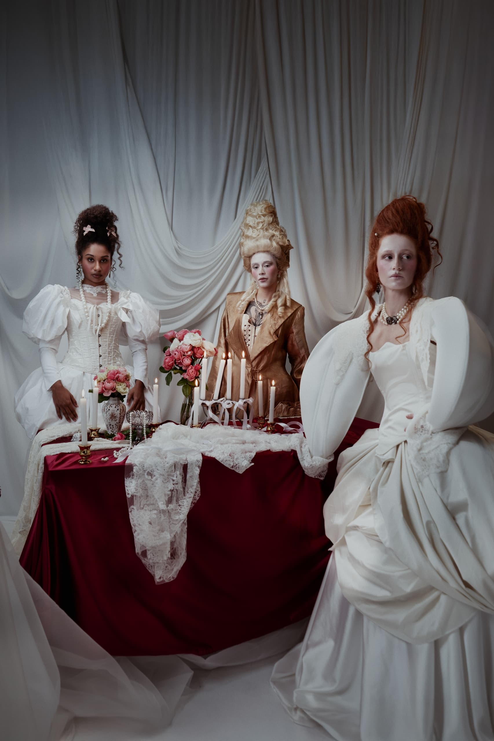 Elegant women in historical gowns at a candlelit table with roses.