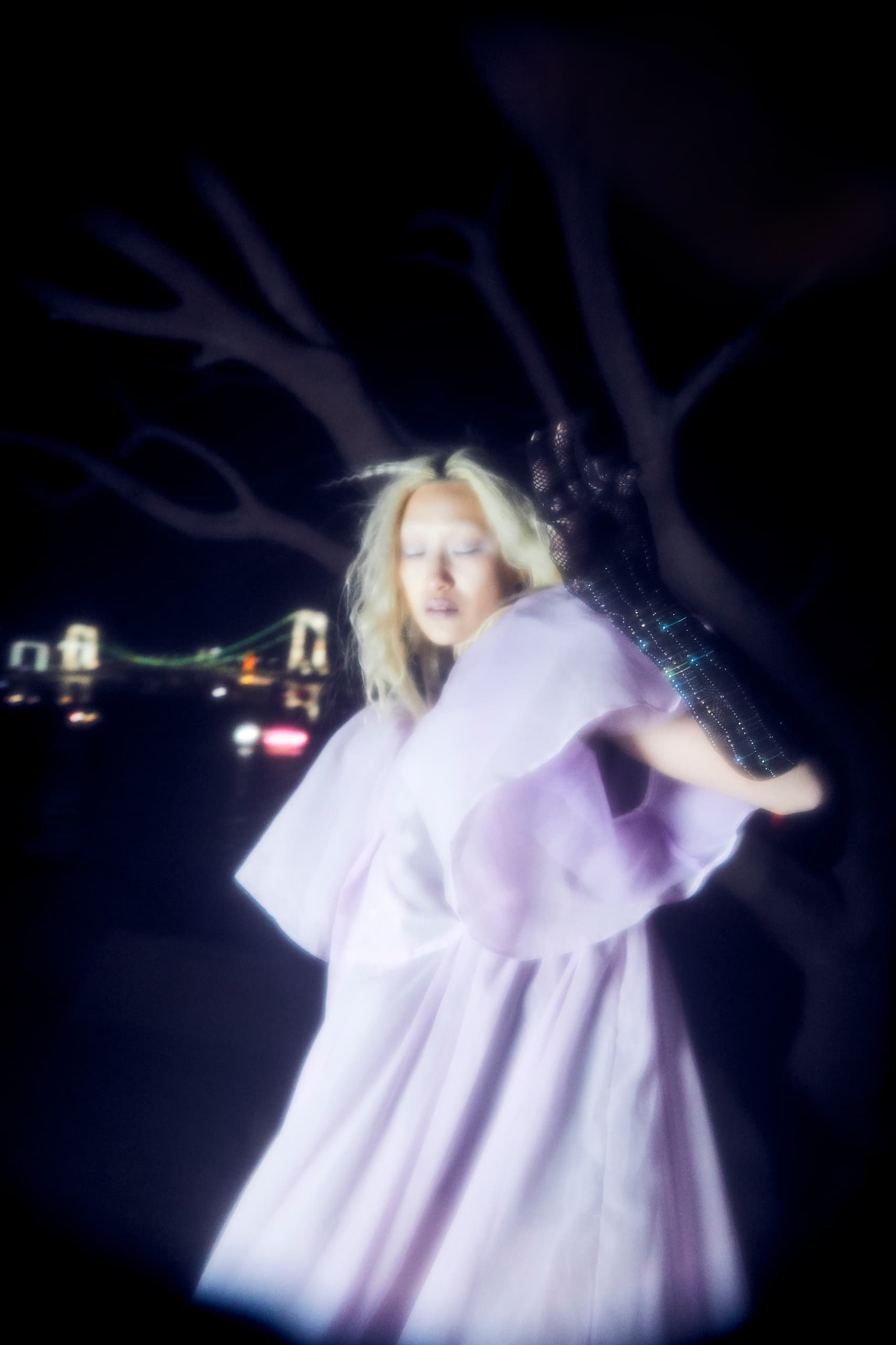 Ethereal woman in lavender dress near illuminated bridge, night fashion scene.