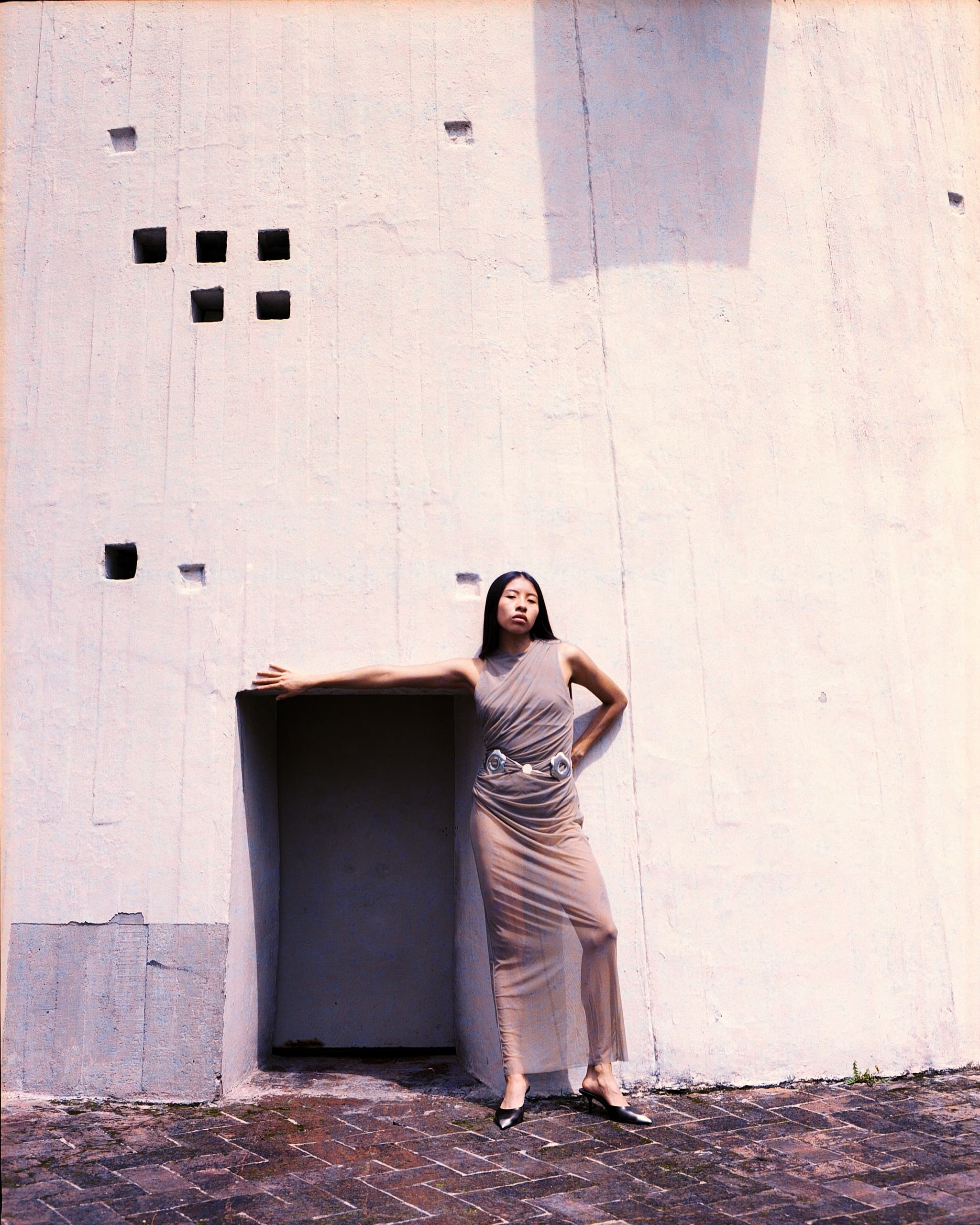Woman in sheer dress standing against a textured wall, embracing minimalist style.