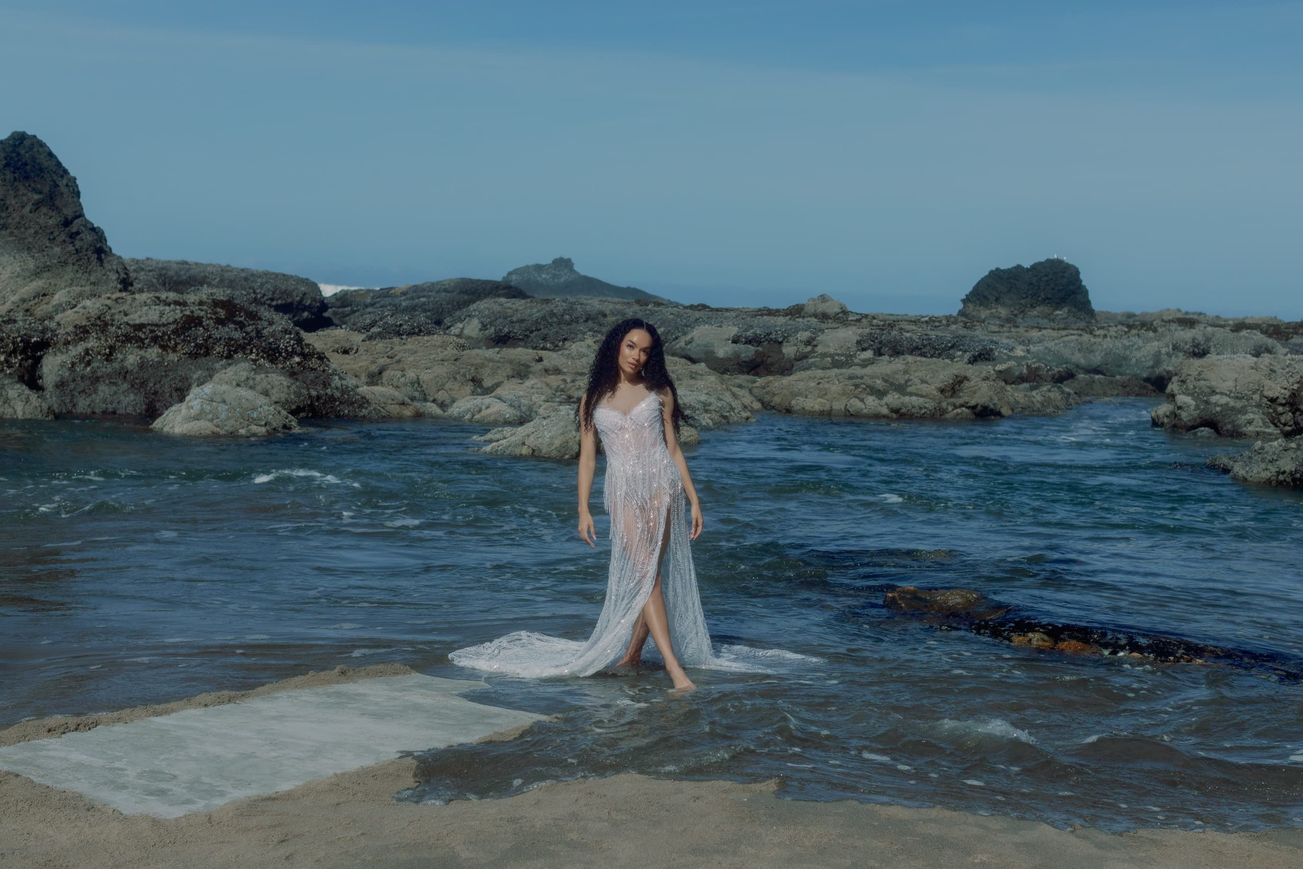 Woman in elegant dress on rocky beach with ocean waves.