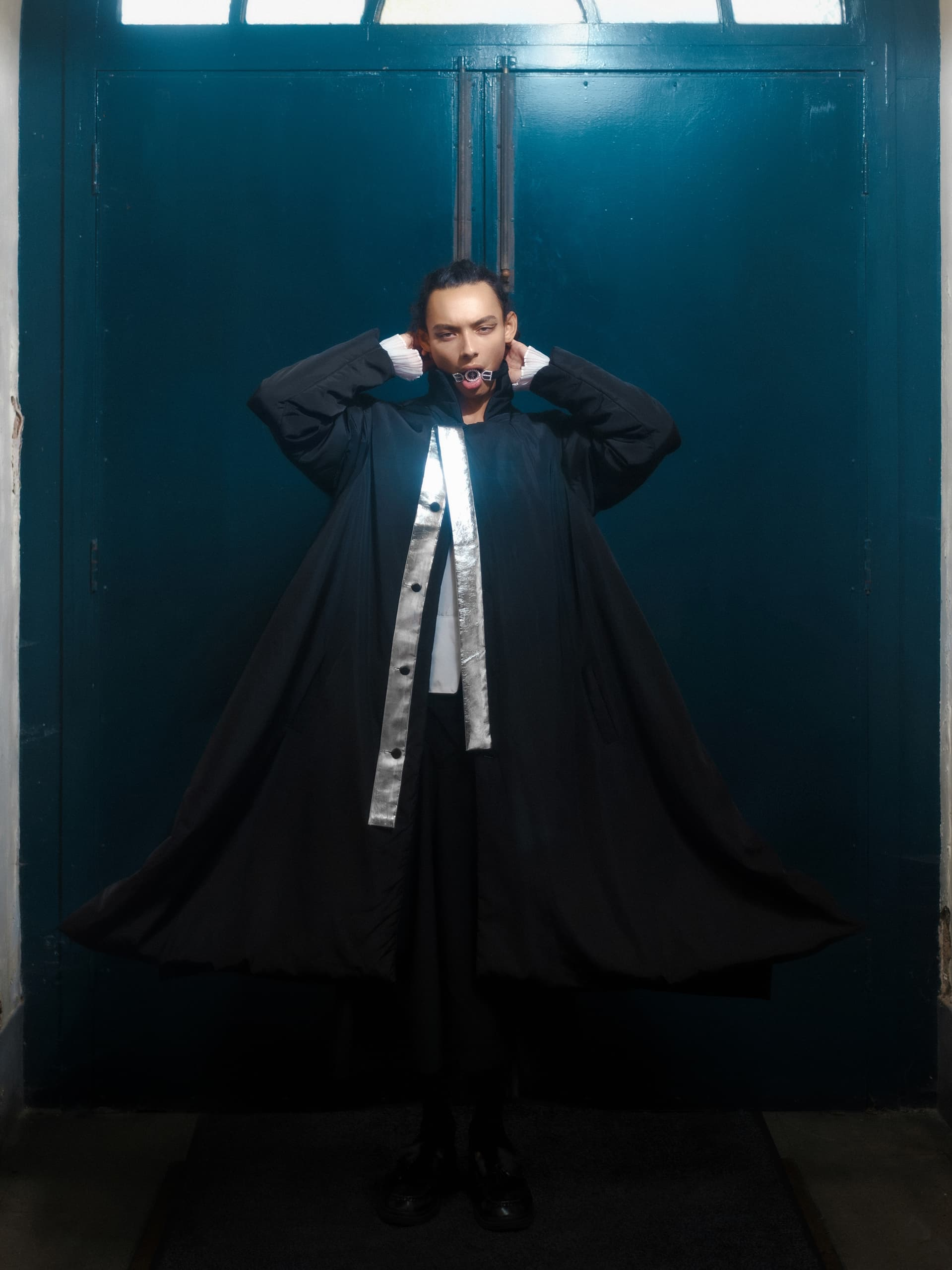 Fashionable person posing in black coat with blue door background.