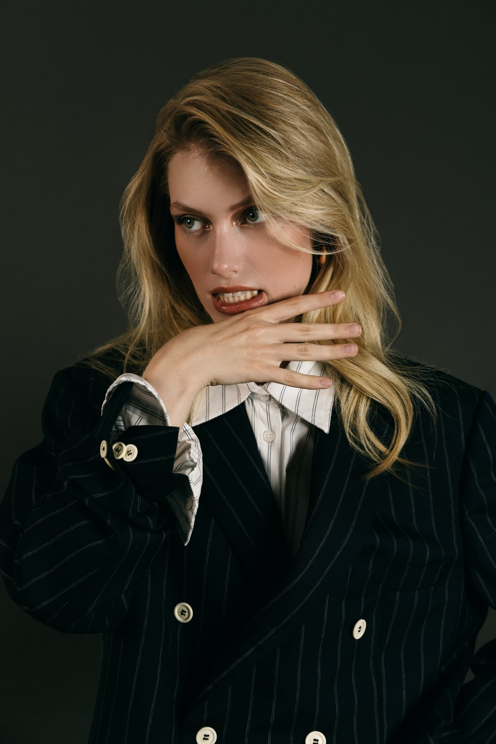 Blonde woman in a pinstripe suit looks thoughtful with hand near her face.