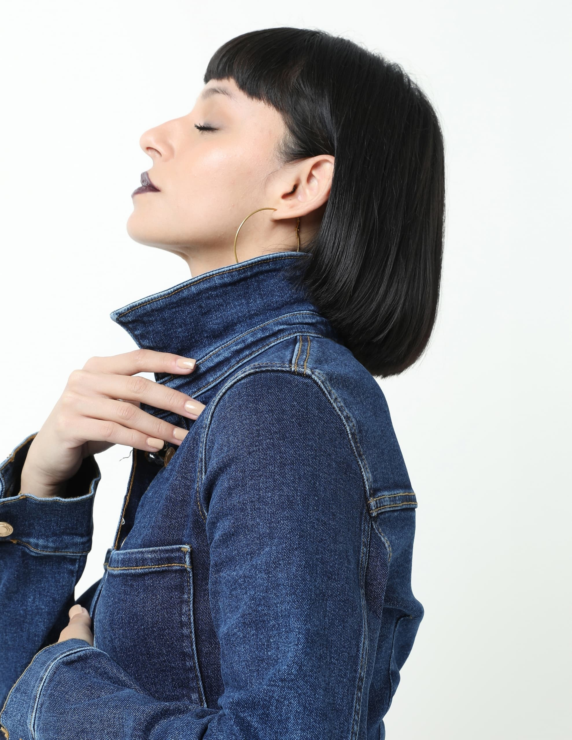 Woman in denim jacket with gold hoop earrings and bob hairstyle, eyes closed.