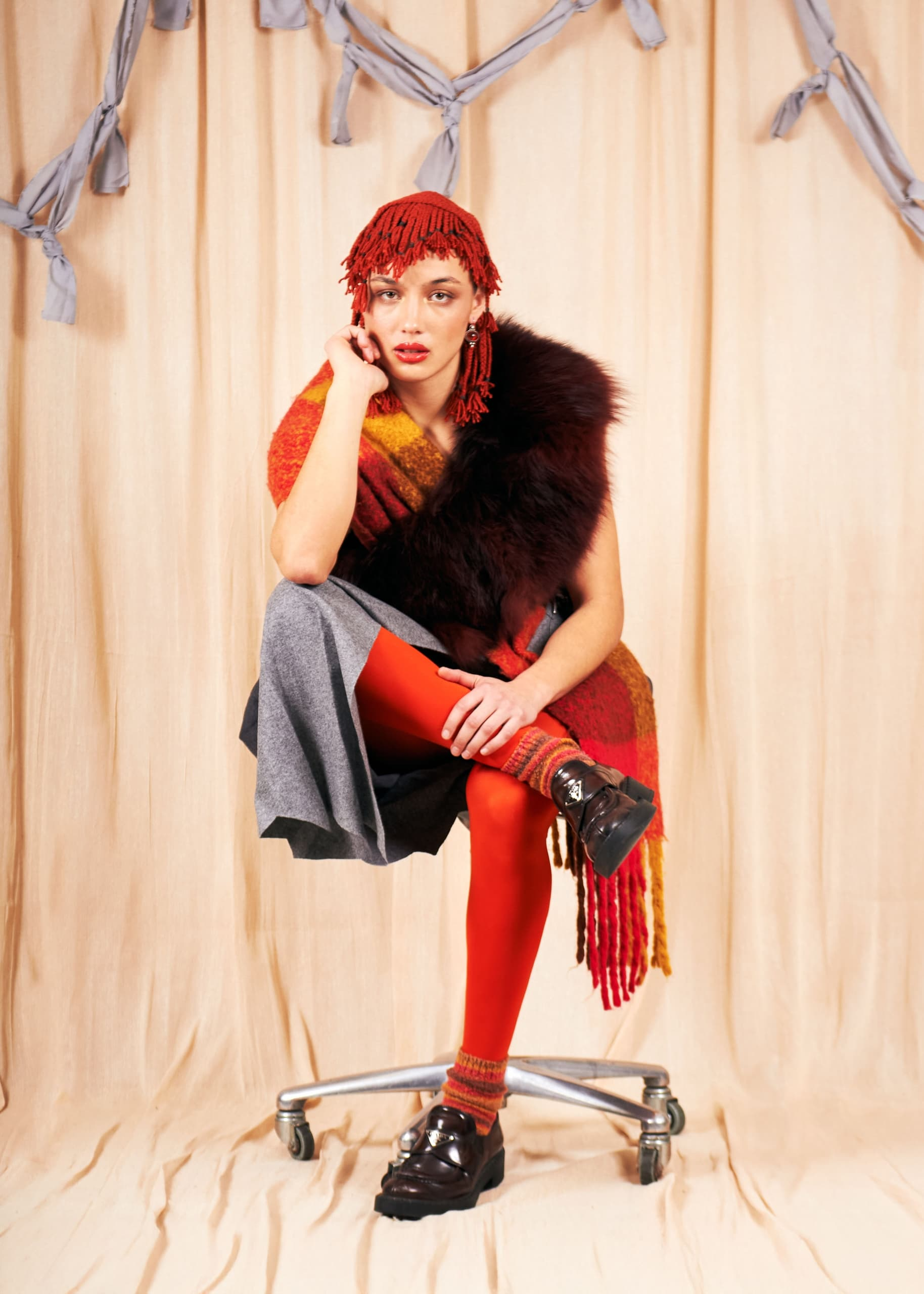 Fashionable person in vibrant ensemble seated against a draped fabric backdrop.