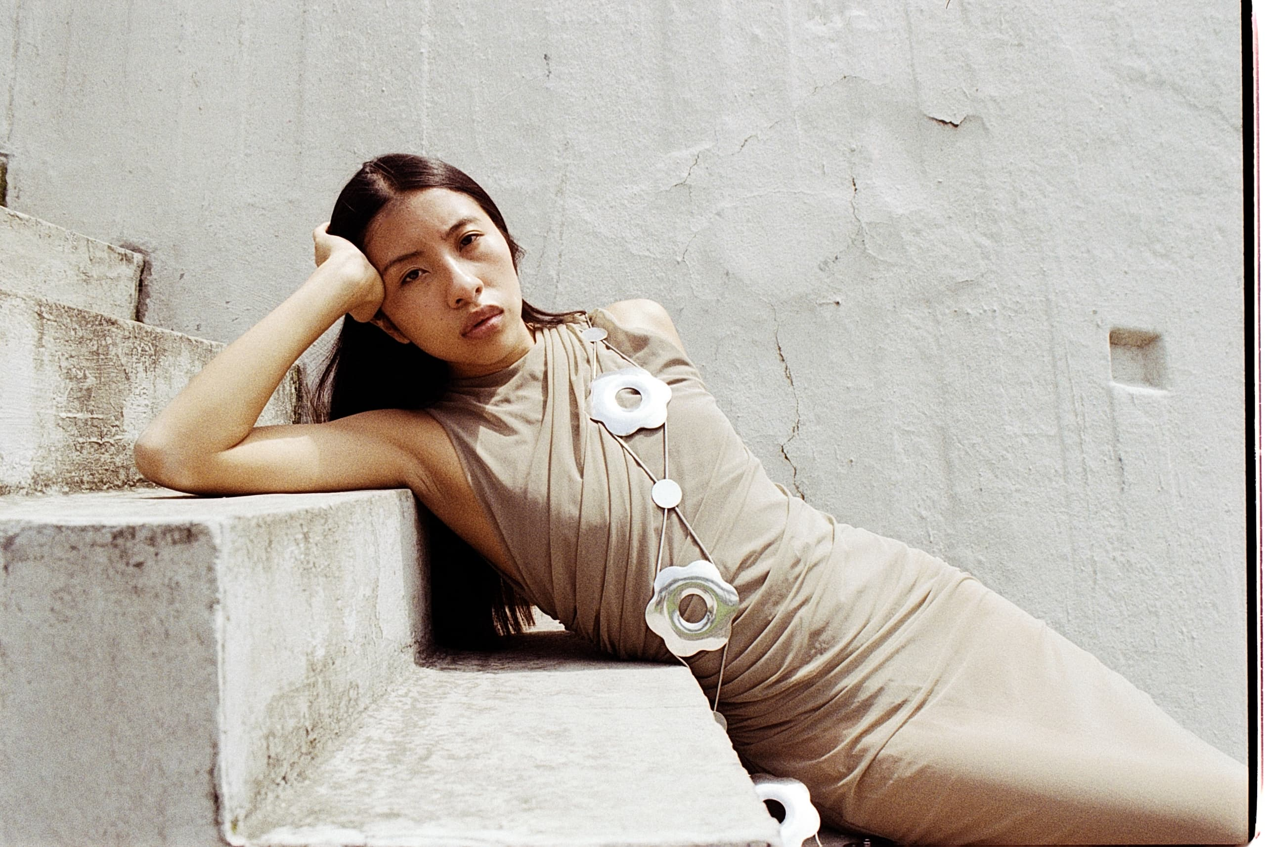 Woman in beige dress with floral necklace, posing on stone steps.