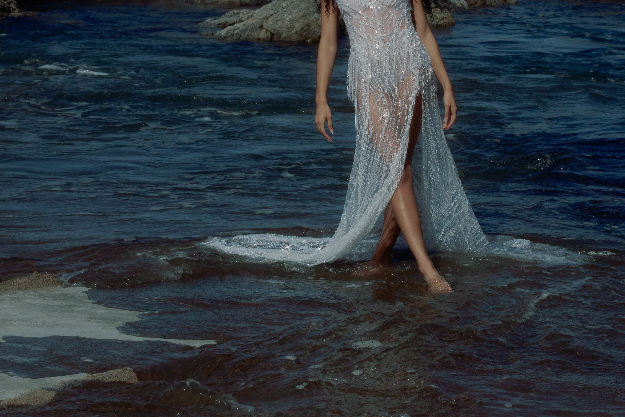 Person in elegant gown wading through water at scenic beach.