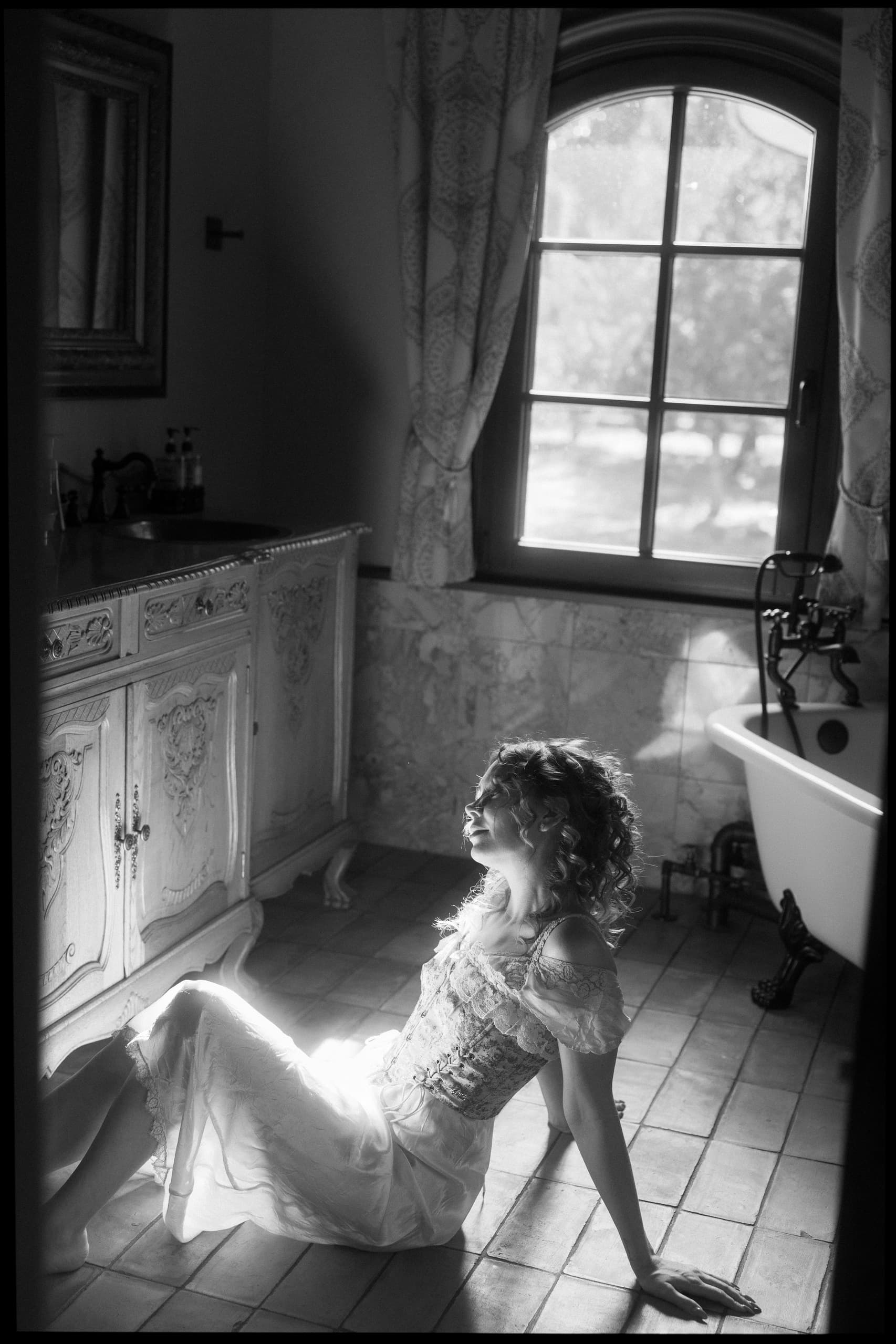 Woman in vintage dress sitting by a sunny window in a classic-style bathroom.