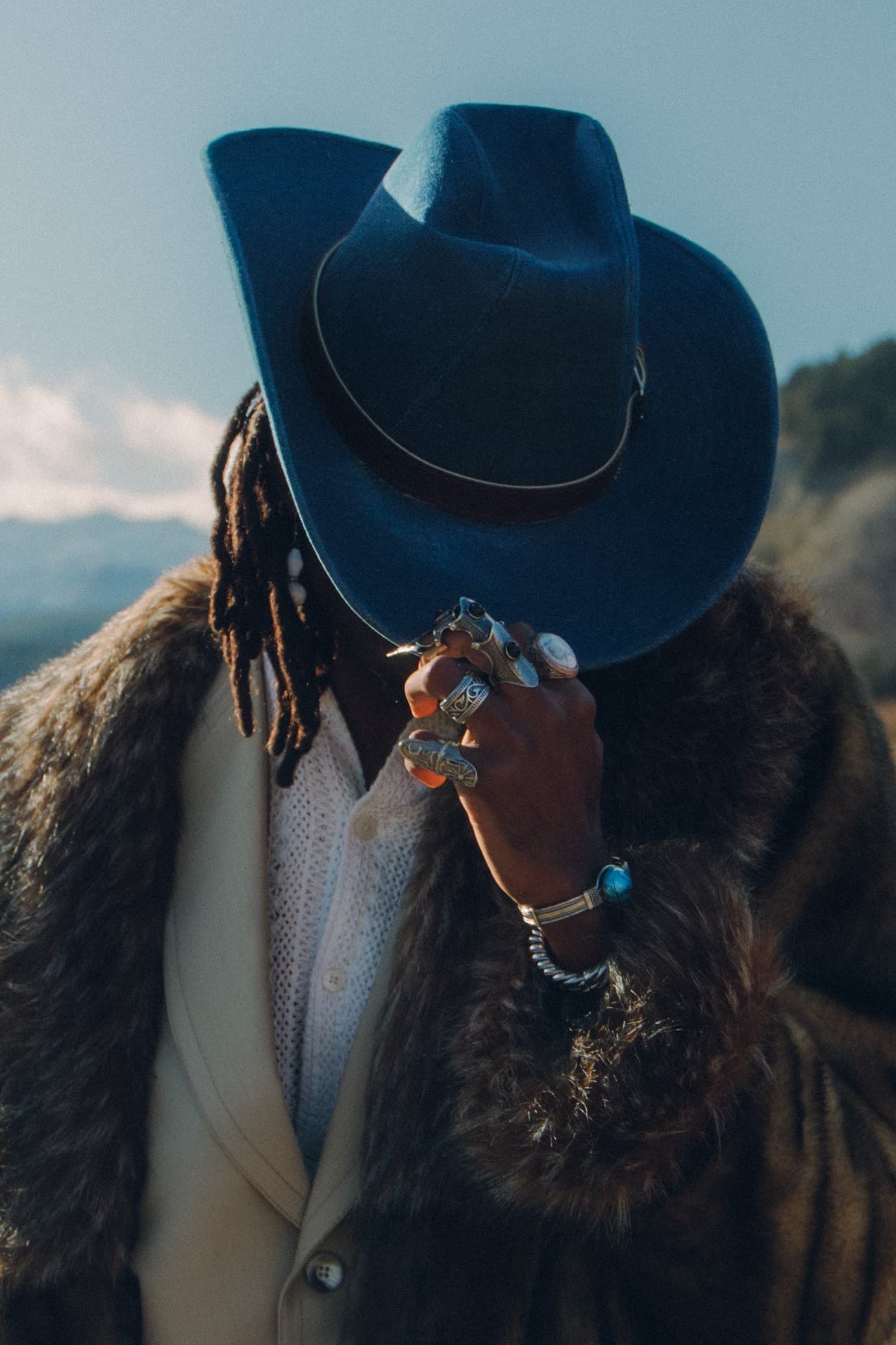 Stylish individual in a blue hat and fur coat, adorned with rings and bracelets.