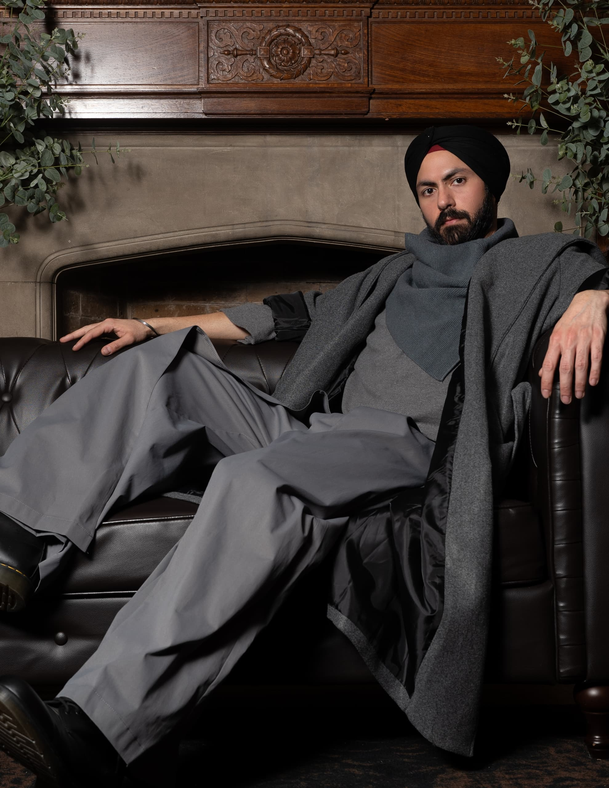 Man in gray attire sits on brown leather couch by ornate fireplace.