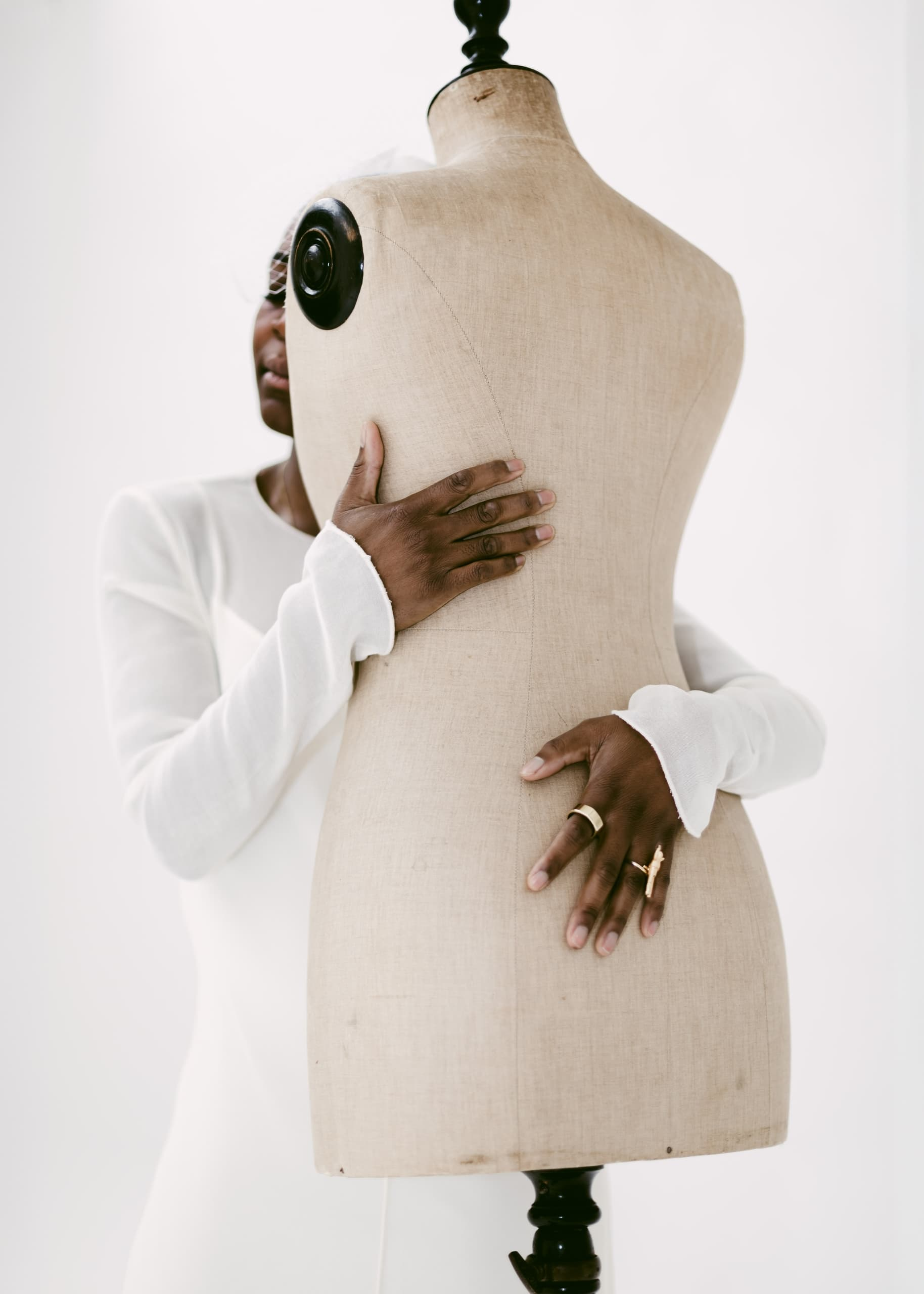 Person embracing dress form mannequin in white setting, wearing stylish rings.