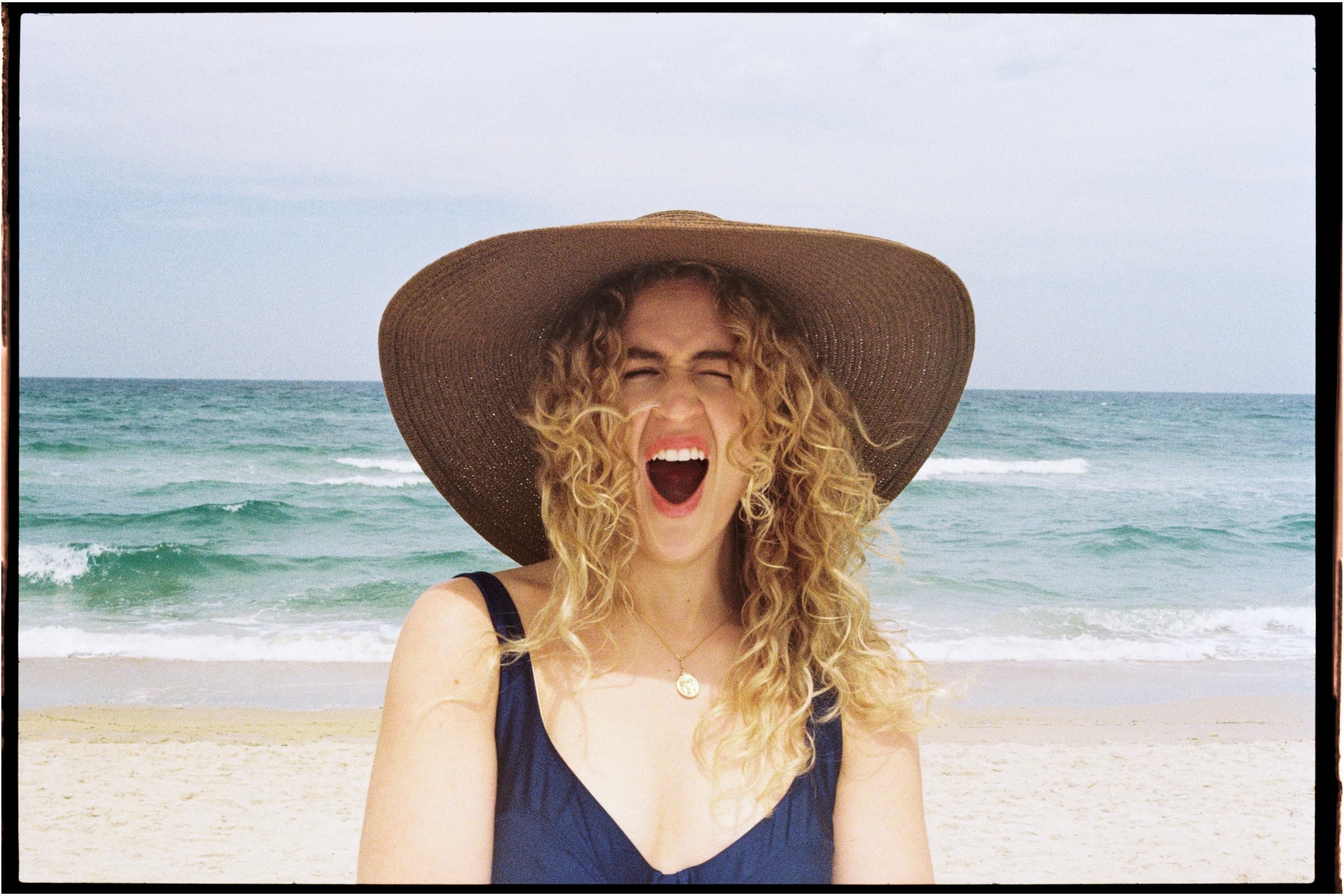 Woman laughing on a beach in a wide-brimmed hat, ocean in the background.