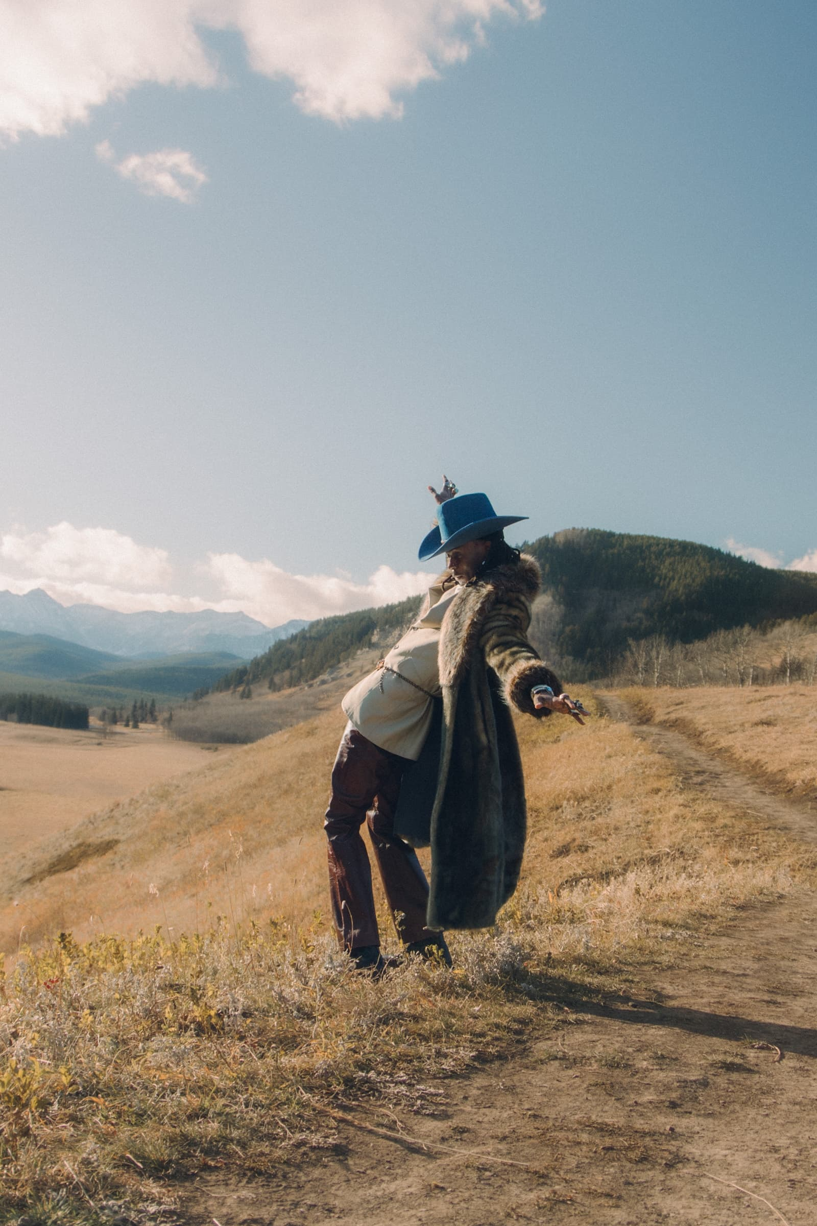Person in fur coat and blue hat posing in scenic mountain landscape.