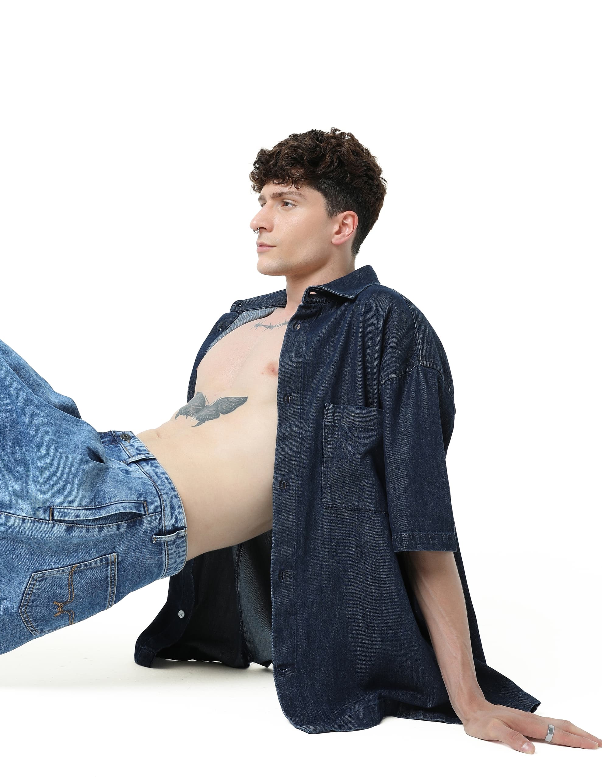 Man in denim outfit poses with a tattoo on his torso against a white background.