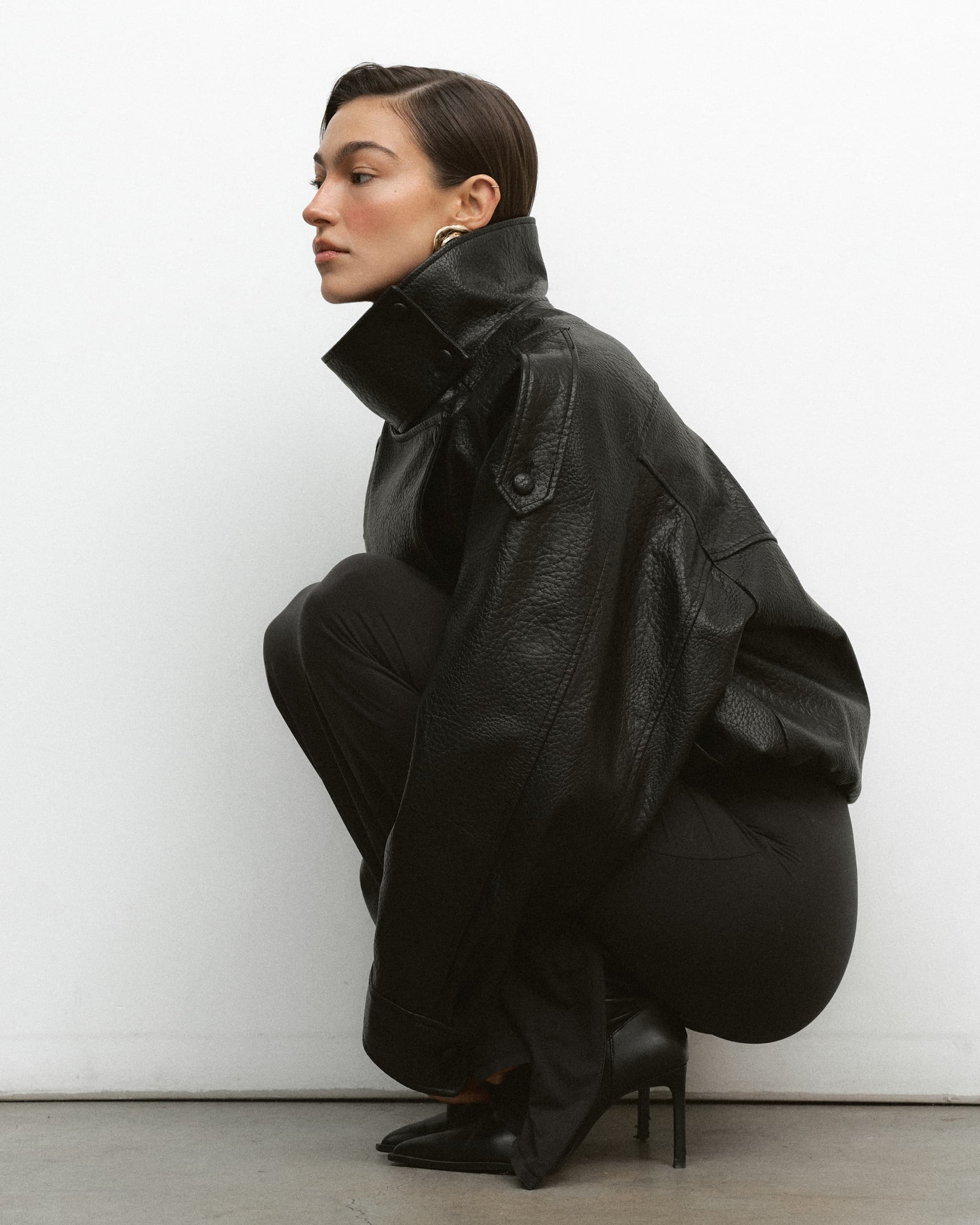 Person squatting in stylish black leather jacket against a white background.