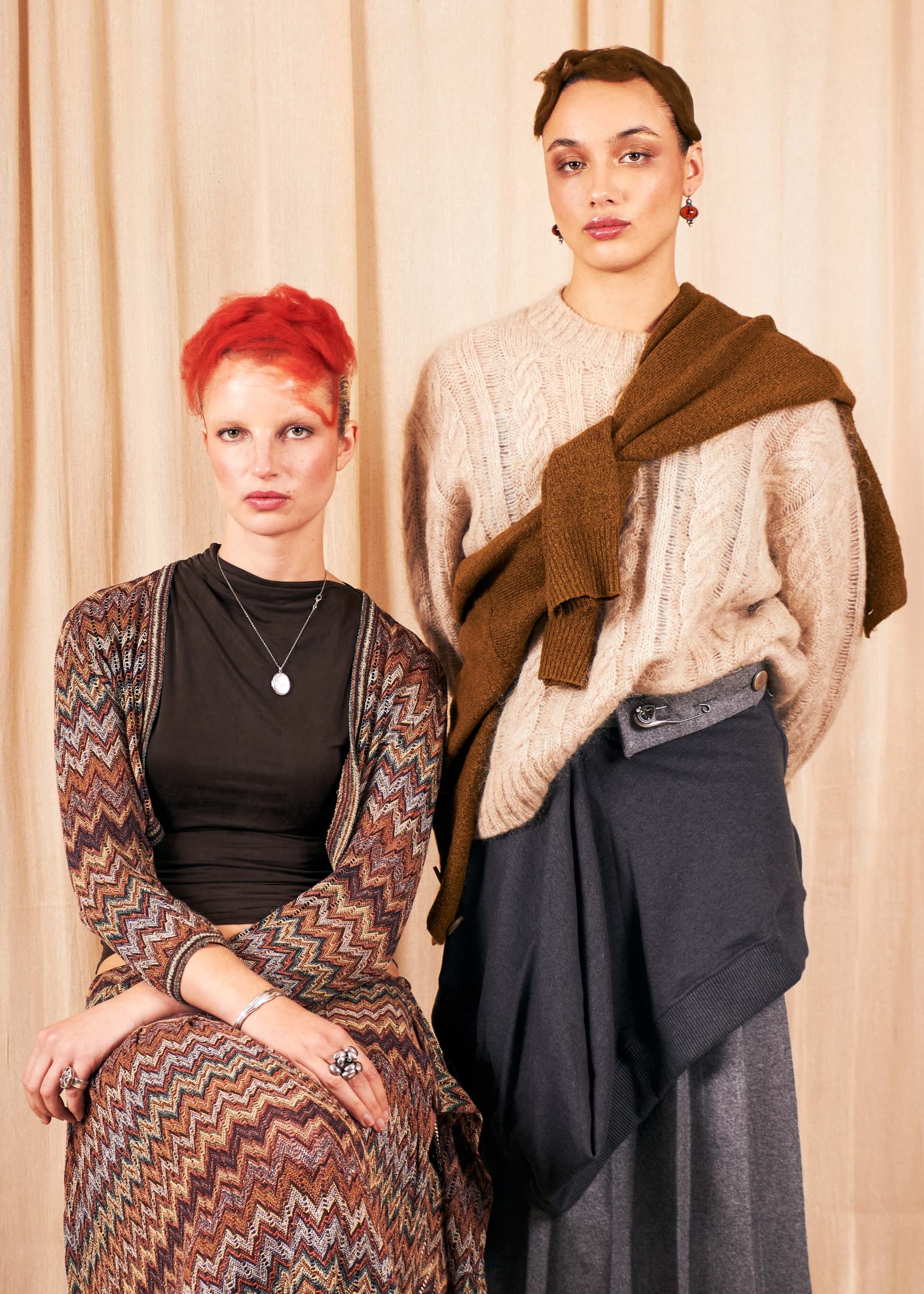 Two models in stylish, layered outfits pose against a beige backdrop. Fashion portrait.