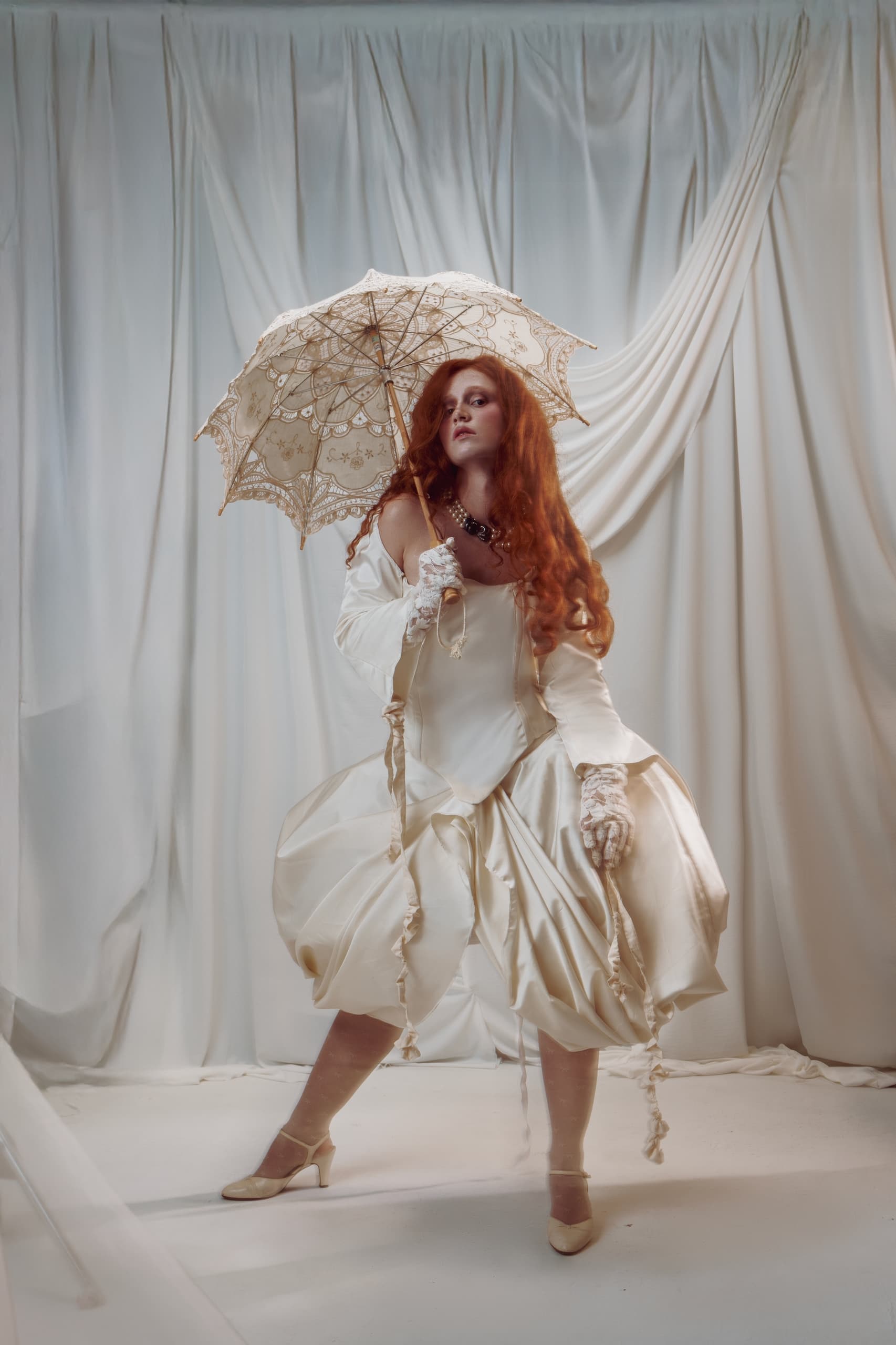 Elegant woman in vintage dress with lace umbrella, posing against draped curtain.