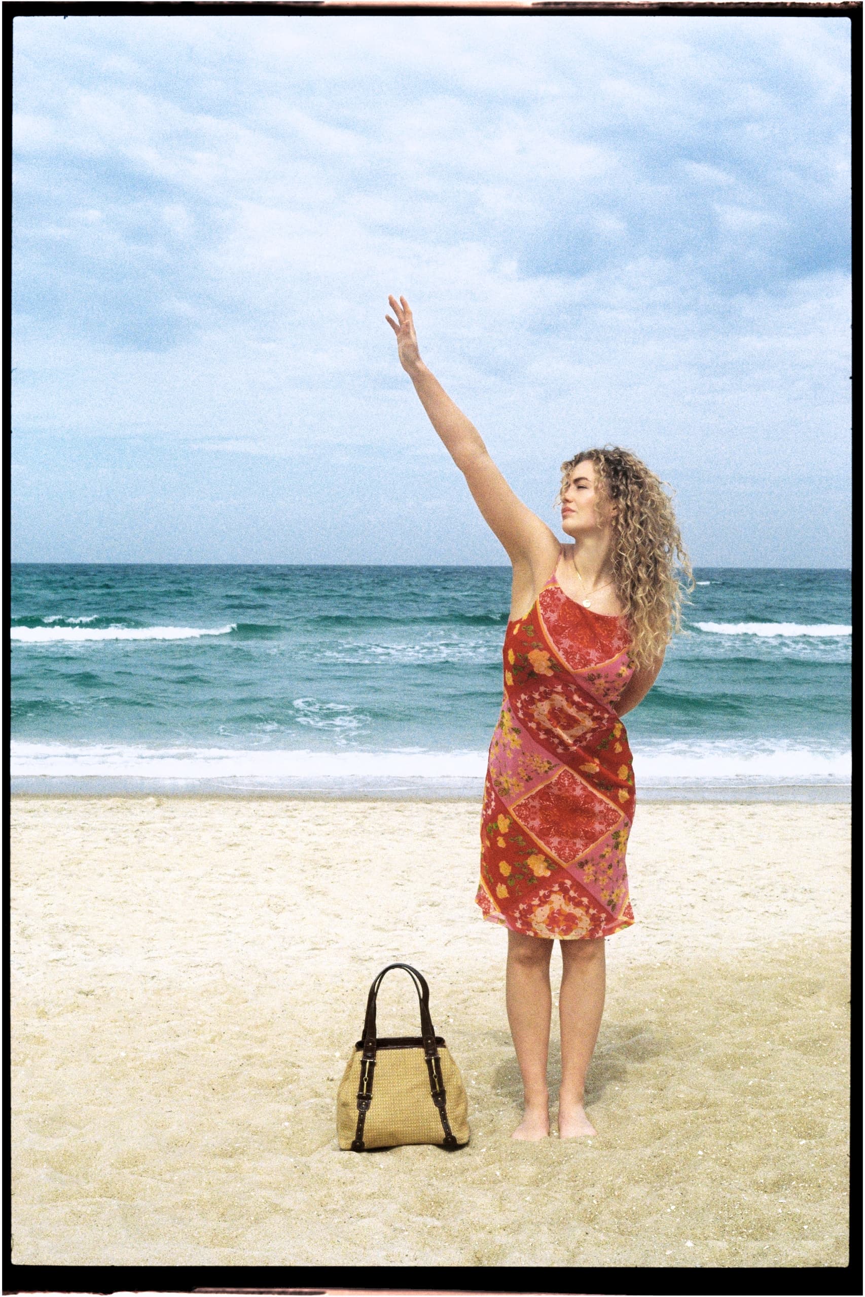Woman in a colorful dress enjoys a sunny beach day, bag by her side, ocean waves behind.