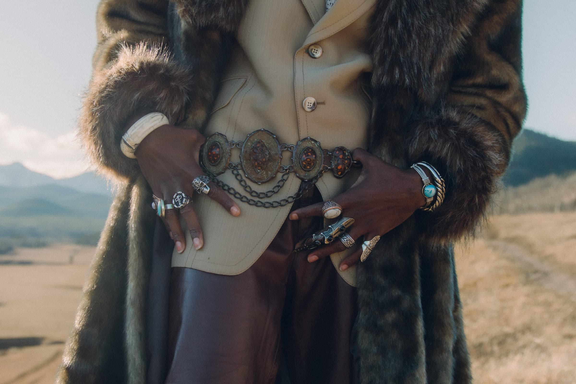Stylish outfit with fur coat, leather belt, and rings in scenic outdoor setting.