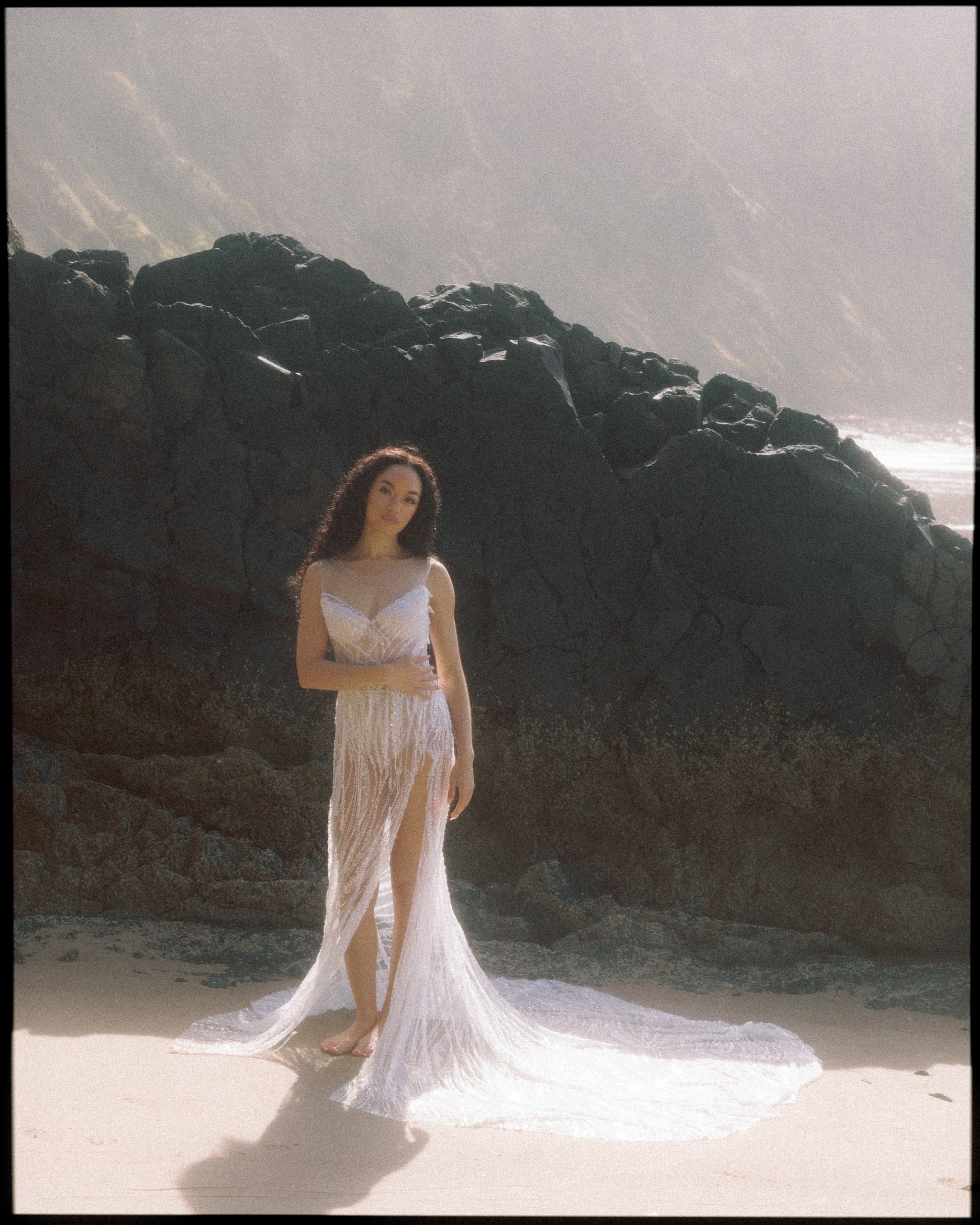 A woman in a flowing white dress stands by rocky cliffs on a sunlit beach.