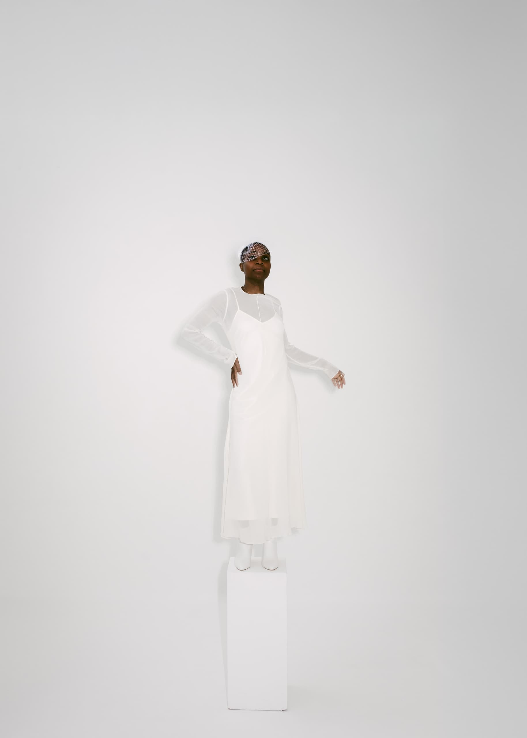 Person in elegant white attire and beaded headpiece standing on a white pedestal.