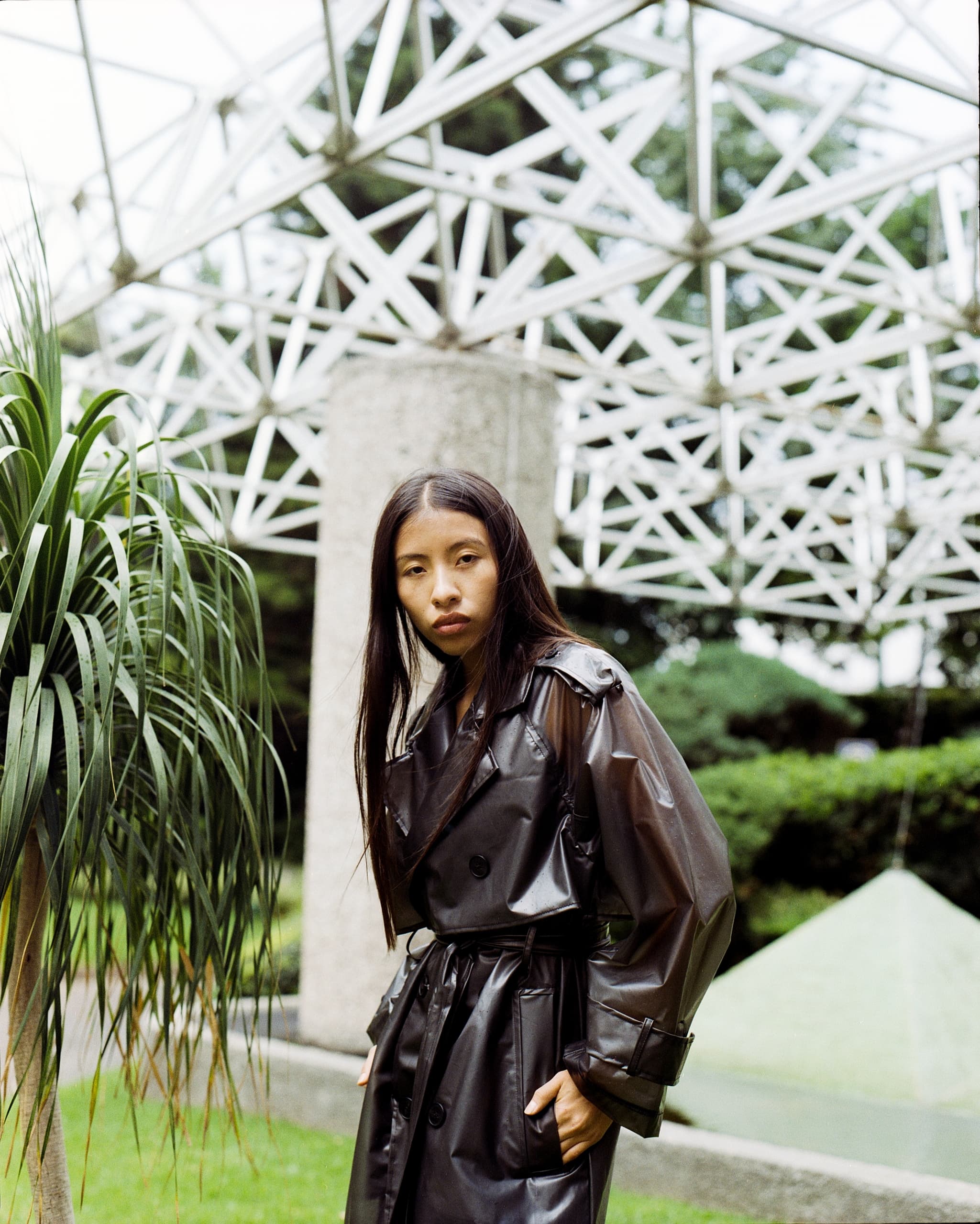 Person in black trench coat outdoors with geometric structure behind.