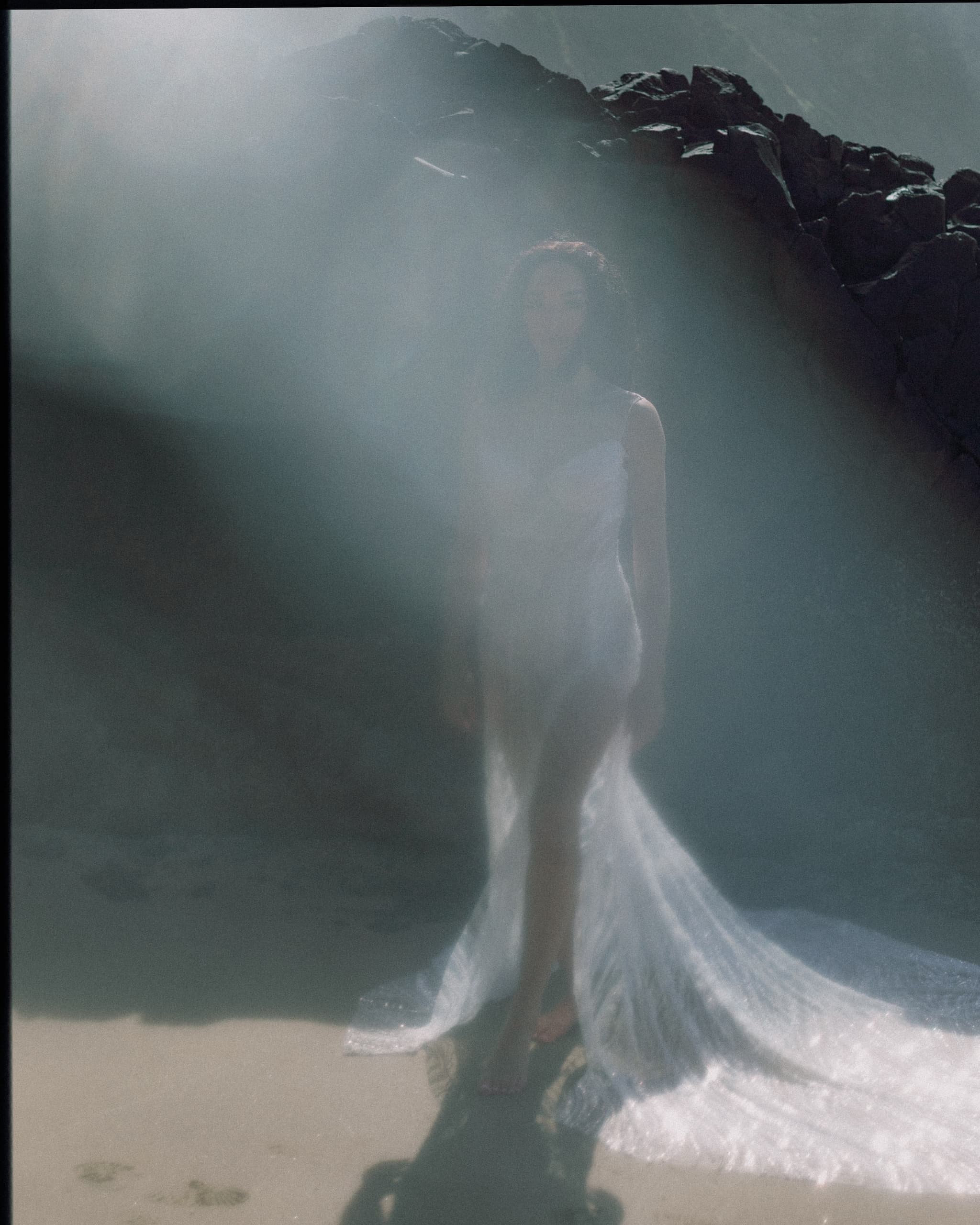 Mysterious woman in ethereal dress standing in sunlight on rocky beach.