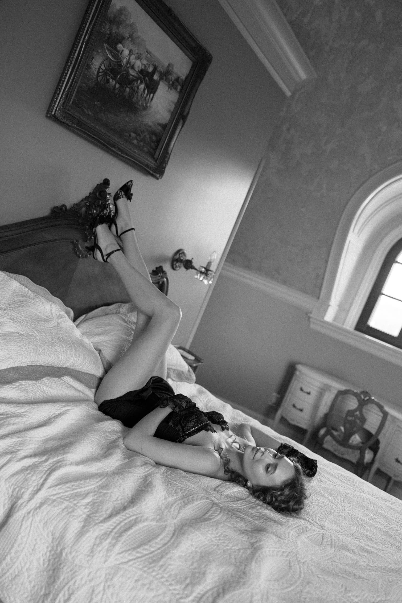 Woman in black outfit relaxing on bed, legs up, elegant bedroom setting.