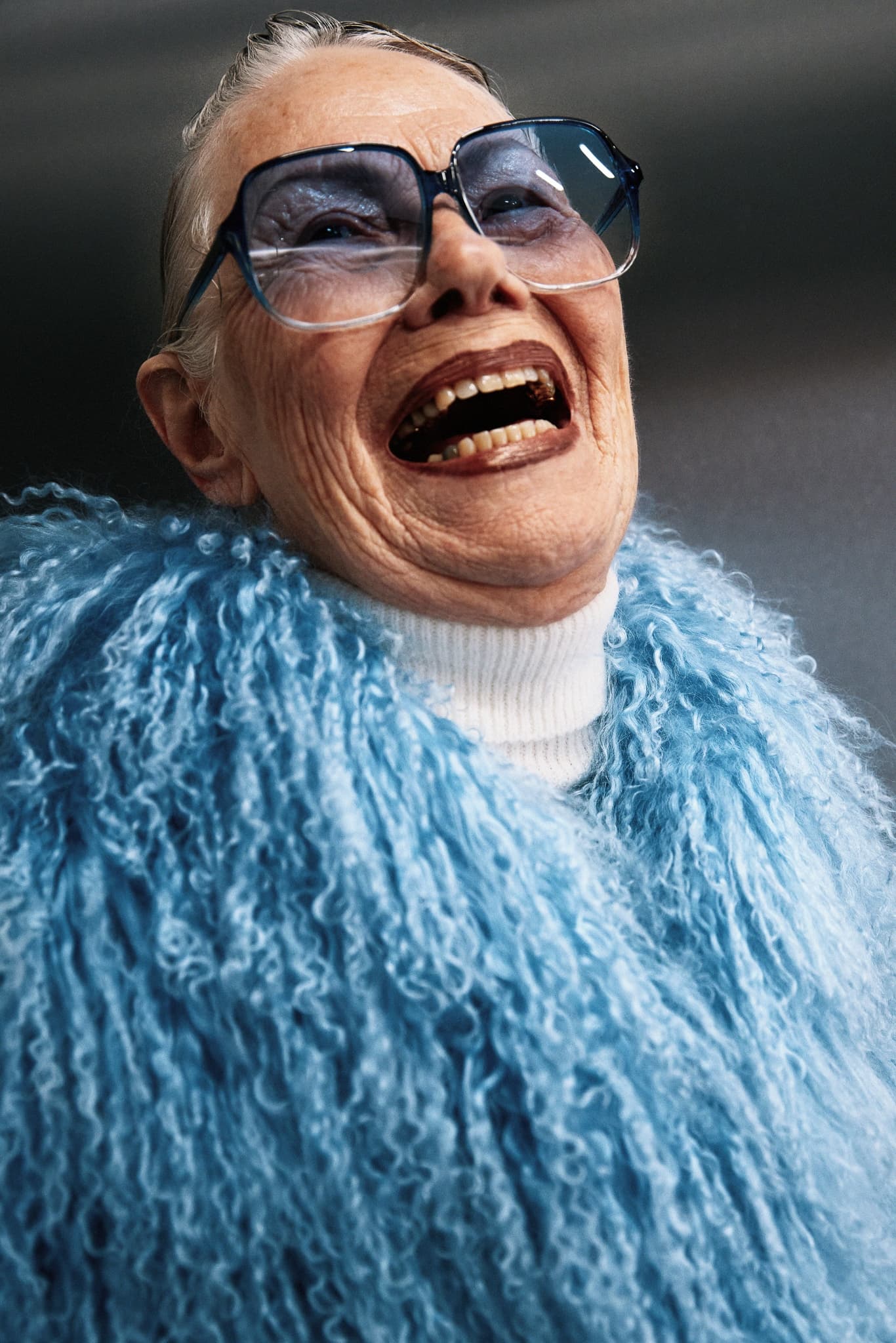 Elderly woman laughing, wearing oversized glasses and a blue textured coat.