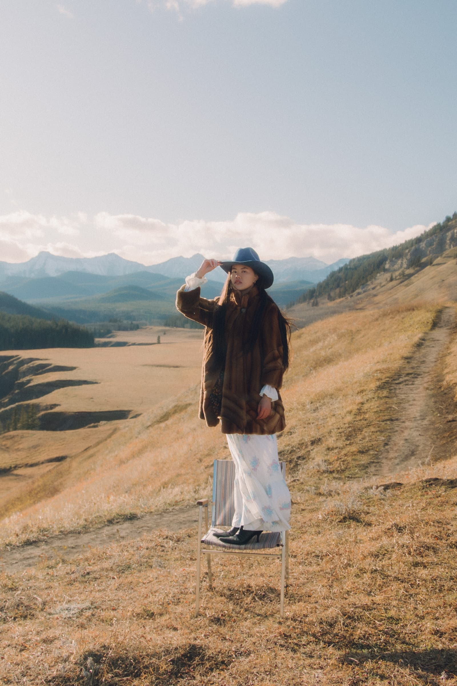 Woman in a hat standing on a chair in a picturesque mountain landscape.