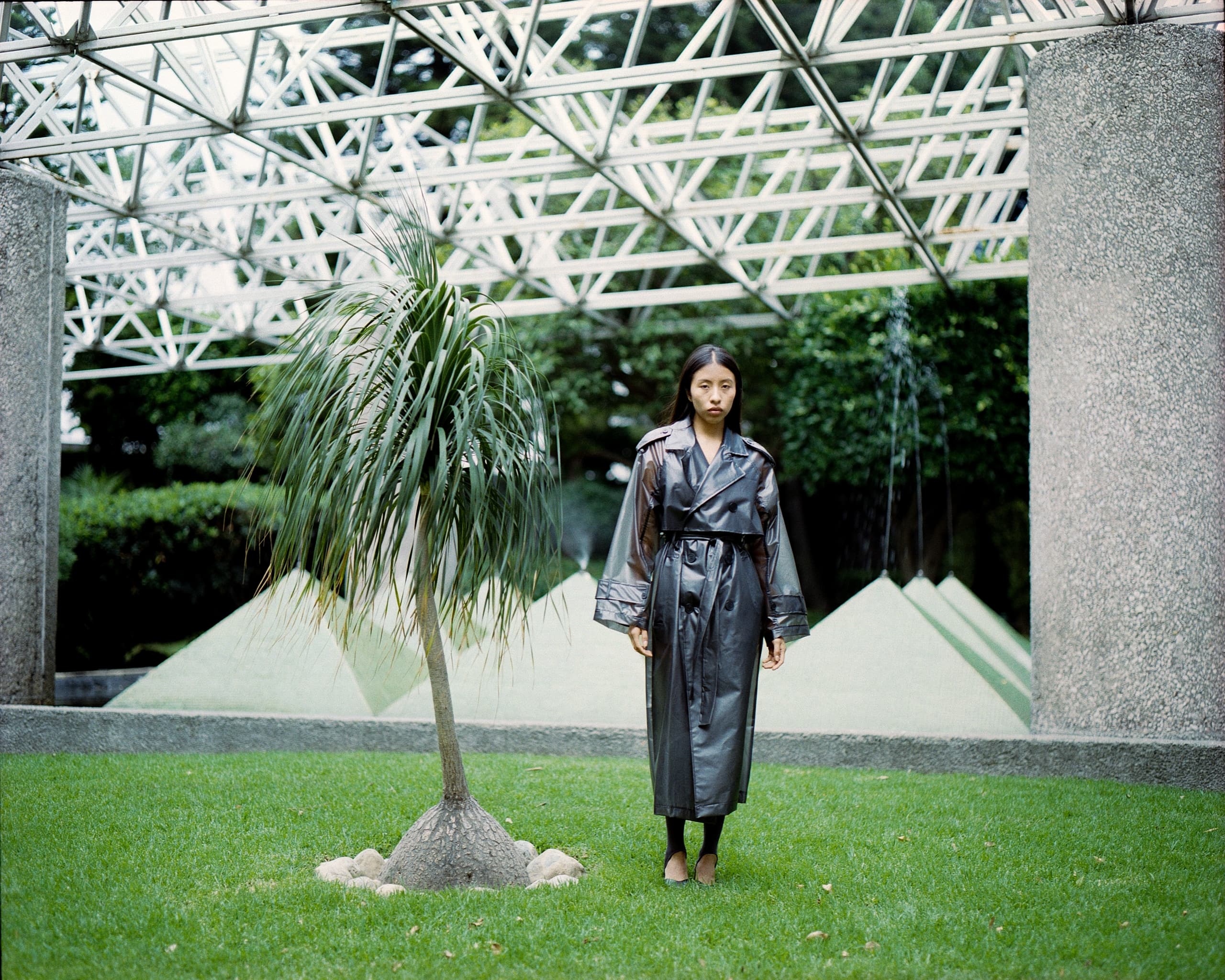 Woman in a black trench coat standing by a tree on a modern outdoor lawn.