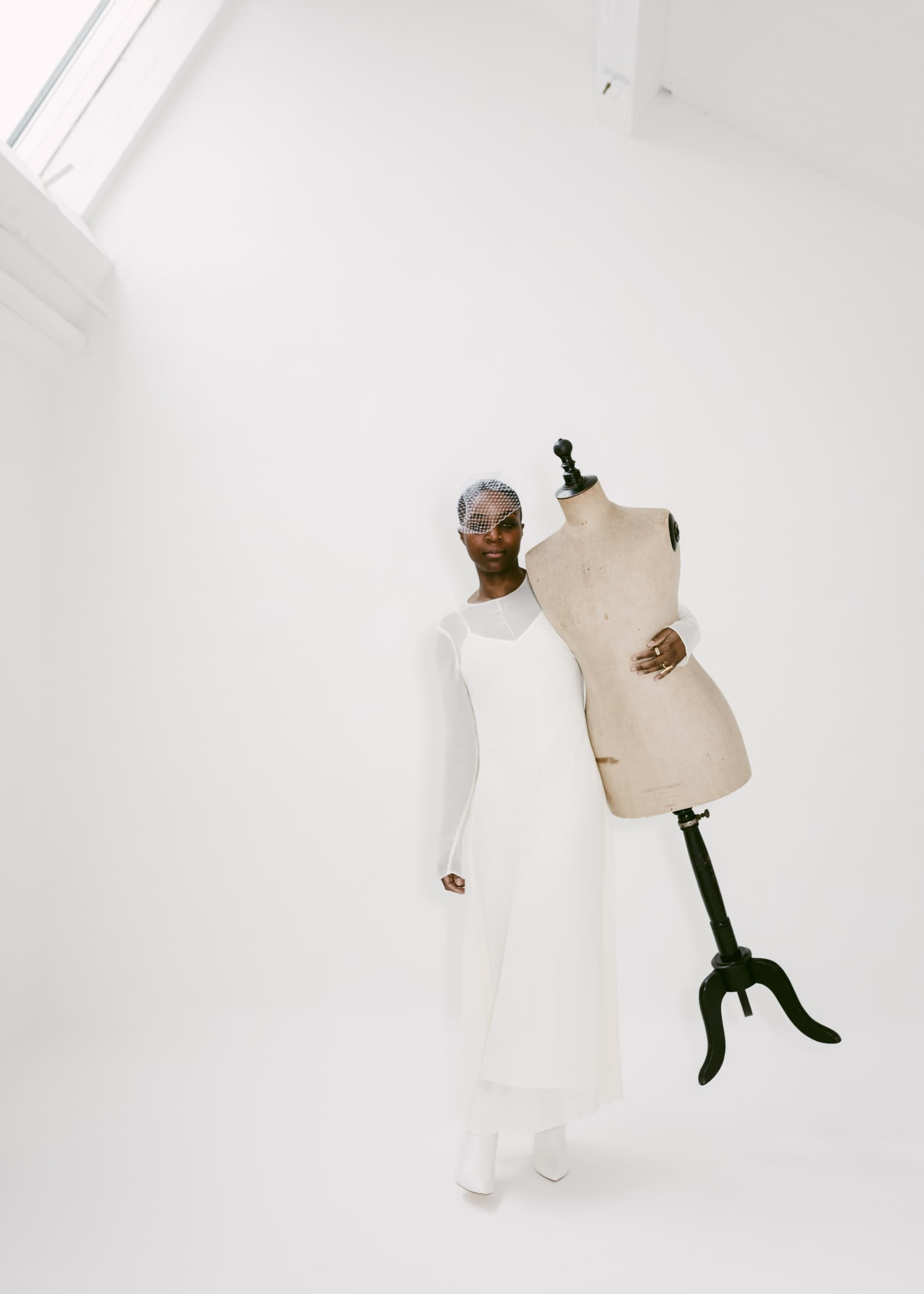 Model in white dress holding mannequin in minimalist room.