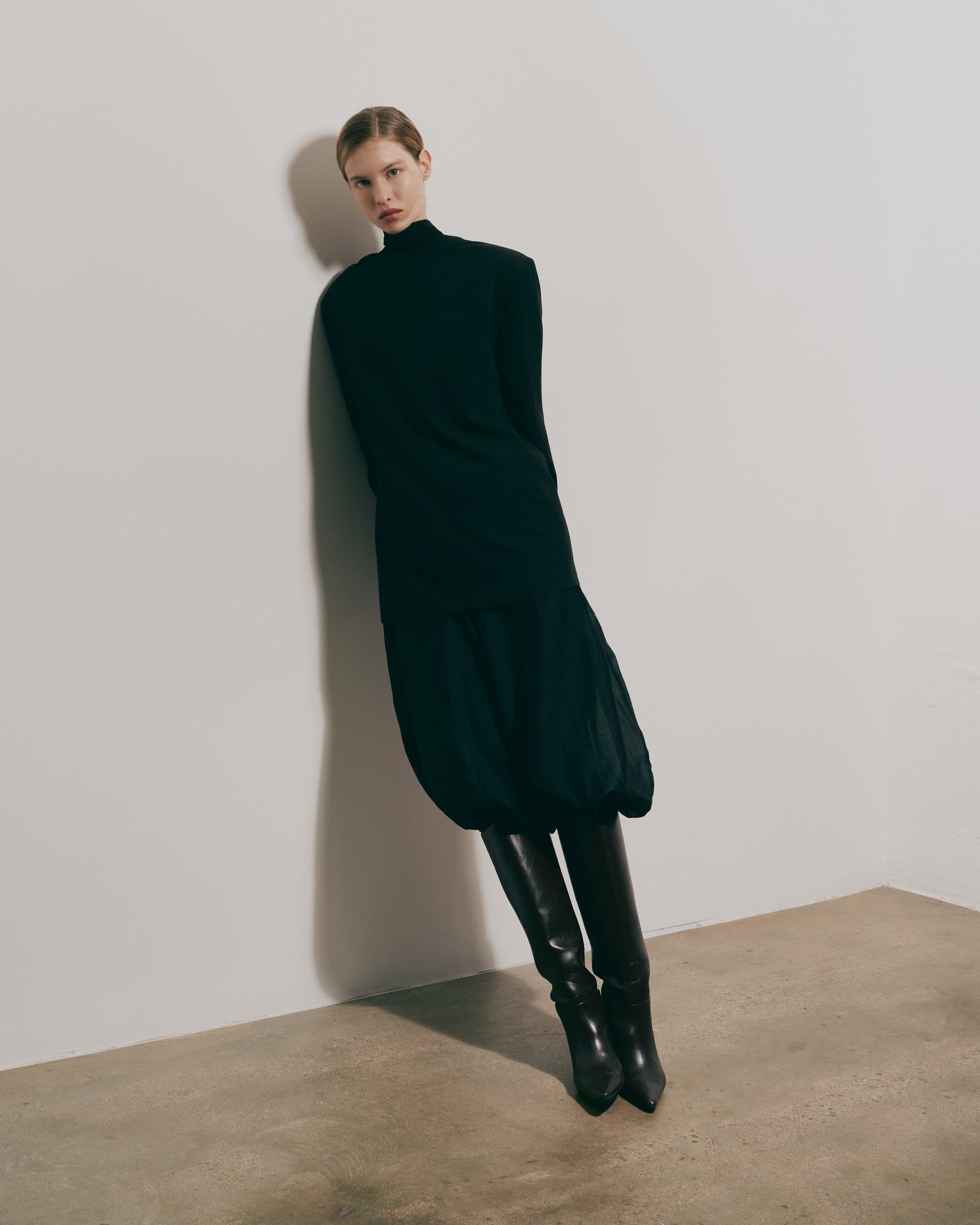 Fashion model in black dress and boots against a white wall.