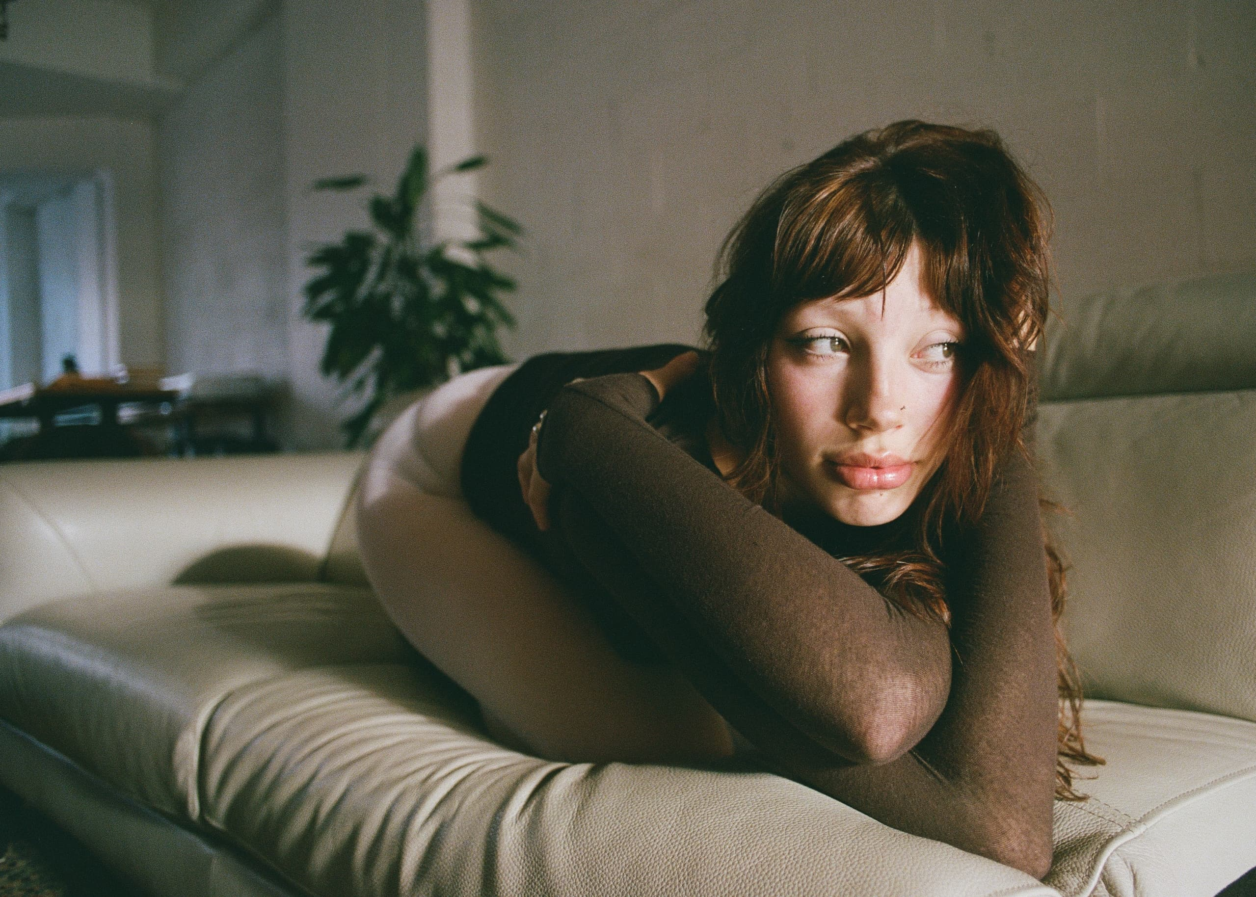Woman in brown top lounging on a sofa, contemplative expression, plant in the background.