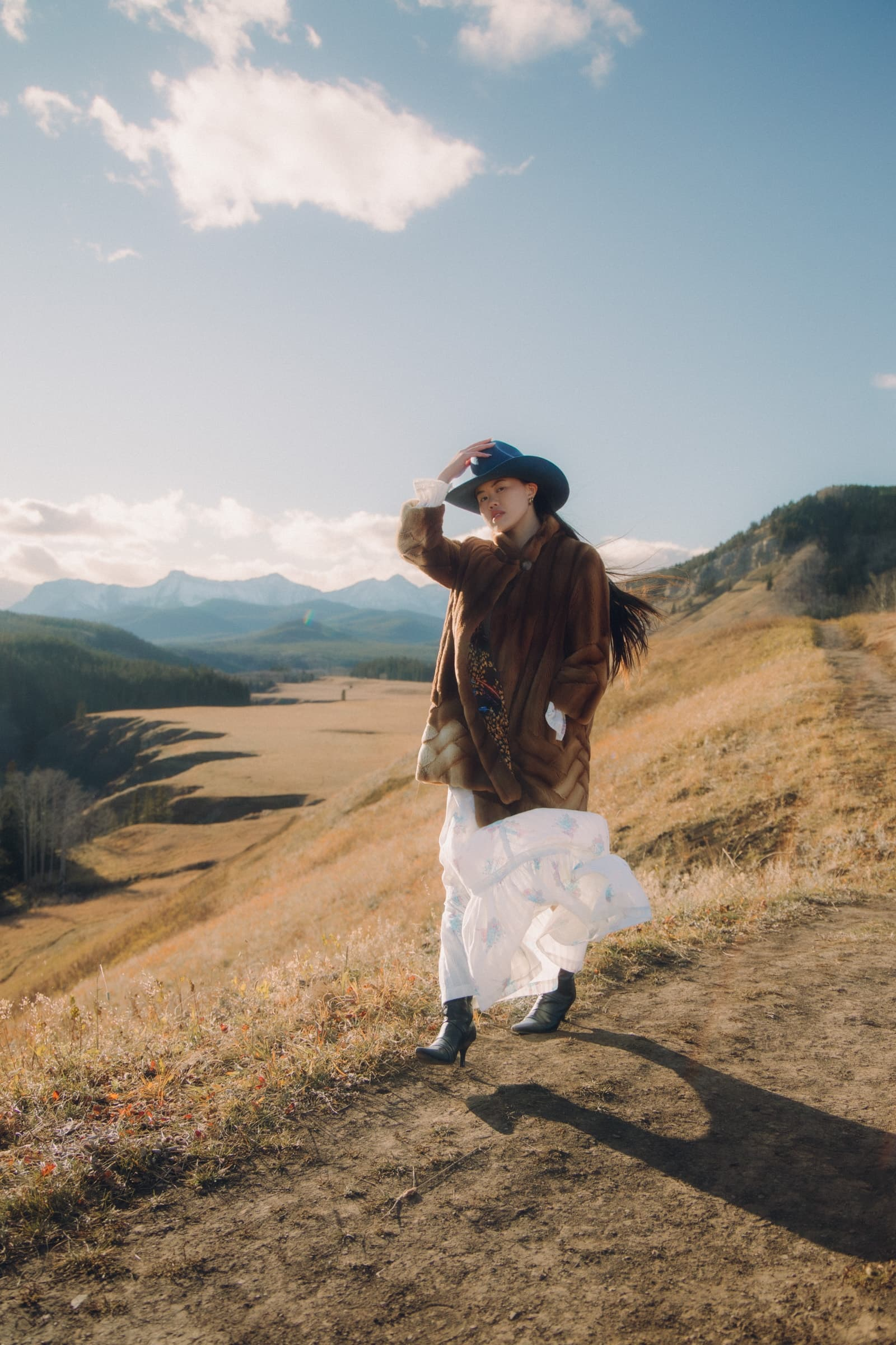 Woman in a hat on a scenic hillside with mountains and blue sky.