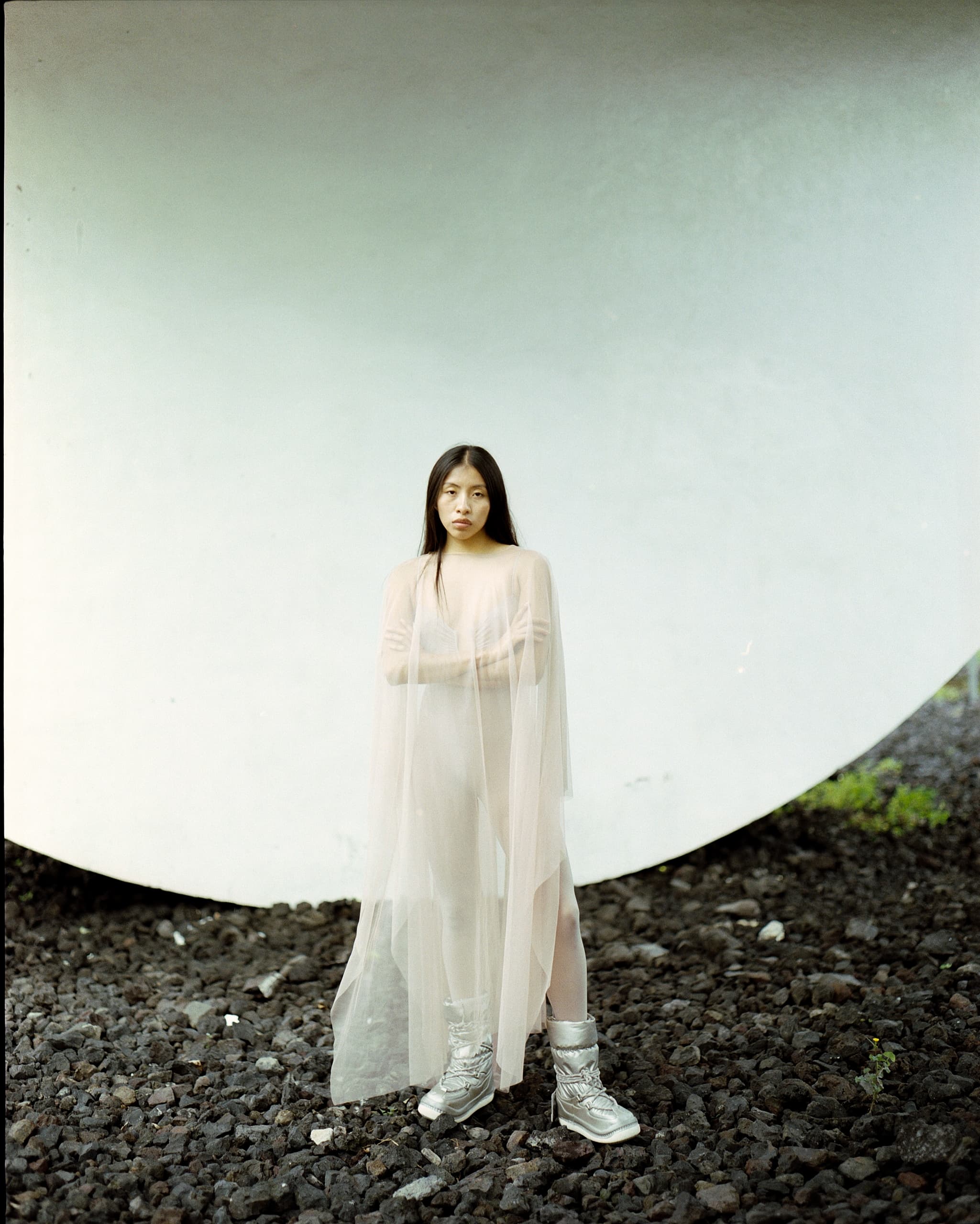 Woman in sheer dress and silver boots standing on rocky ground, minimalist backdrop.