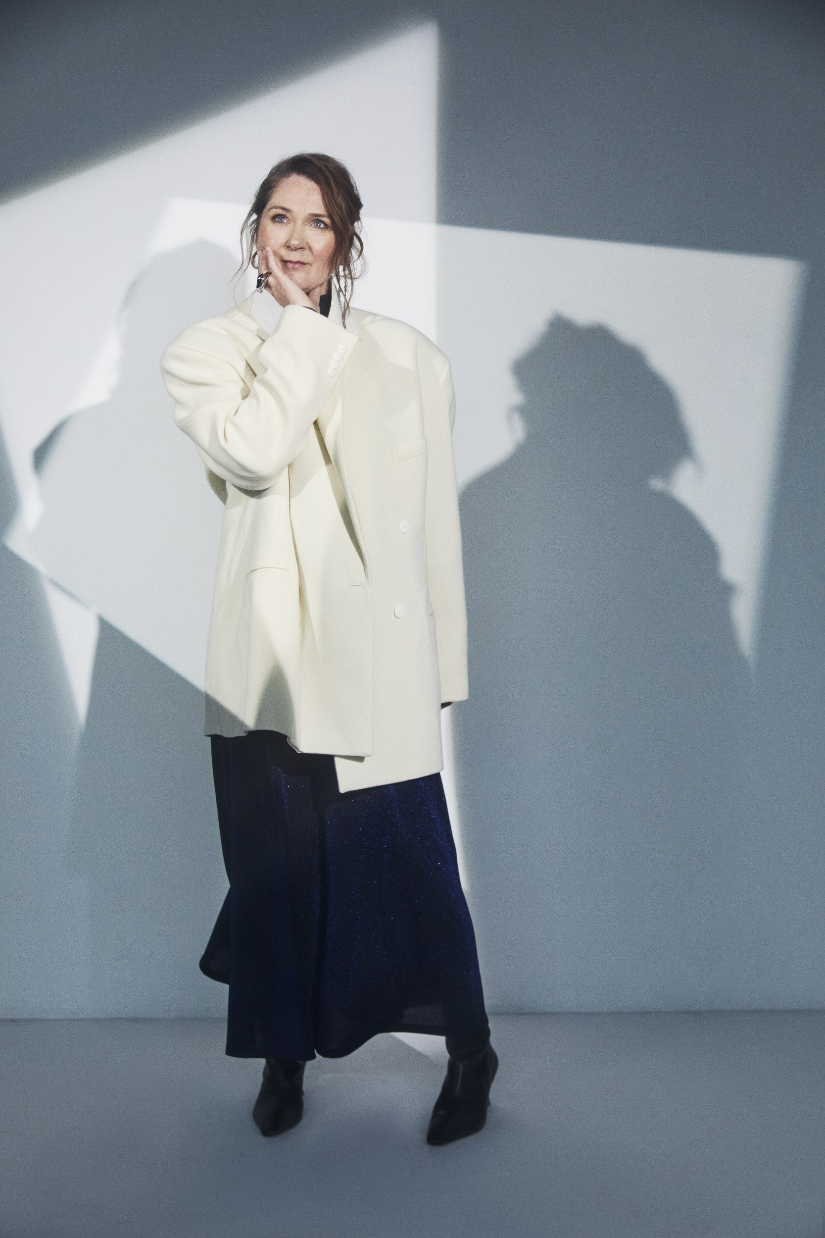 Woman in oversized white blazer, blue skirt; geometric shadows on wall.