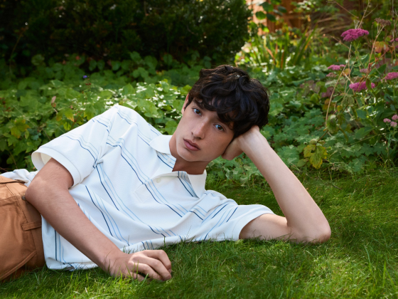Person relaxing on grass in garden, wearing a striped polo and brown pants.