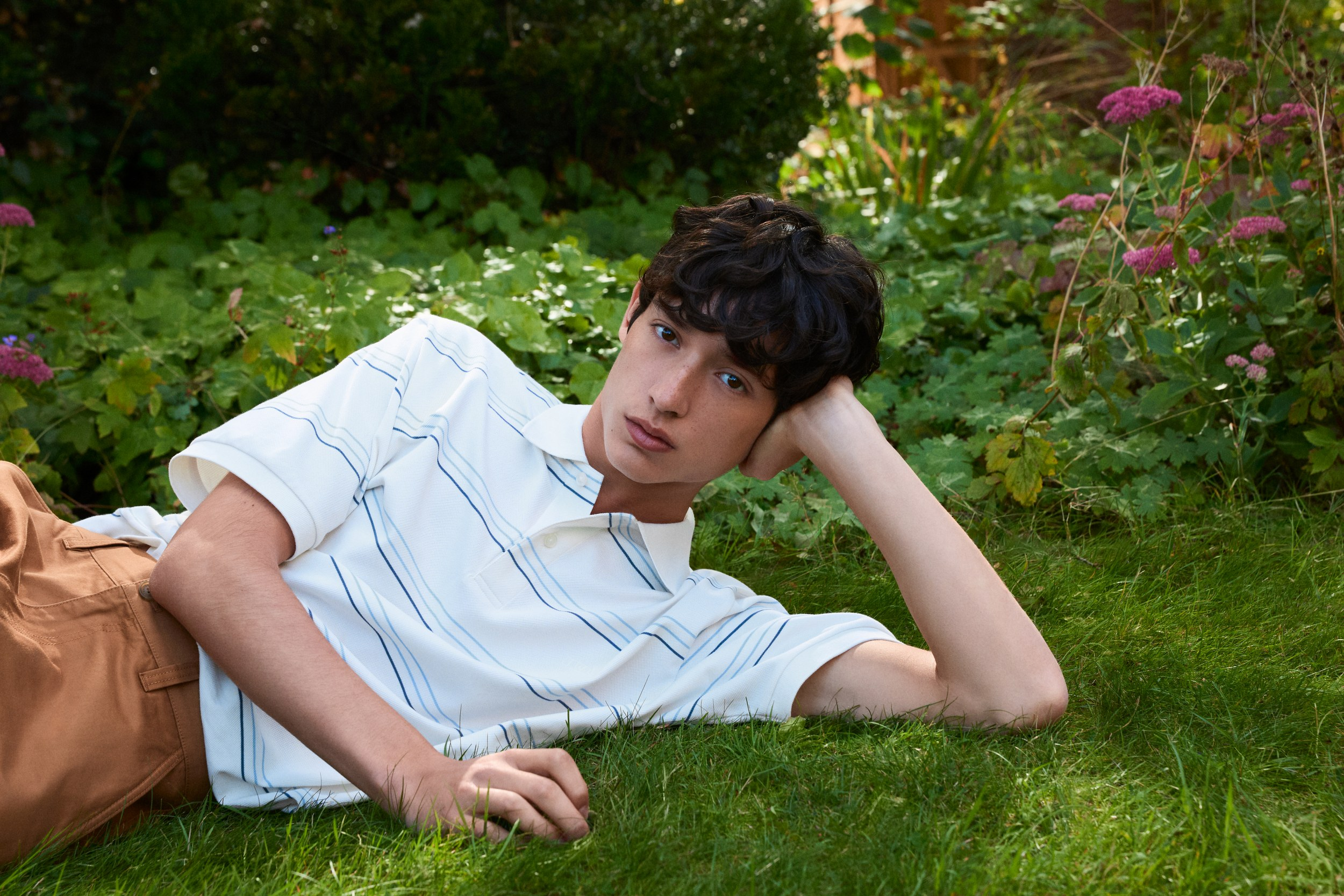 Person relaxing on grass in garden, wearing a striped polo and brown pants.